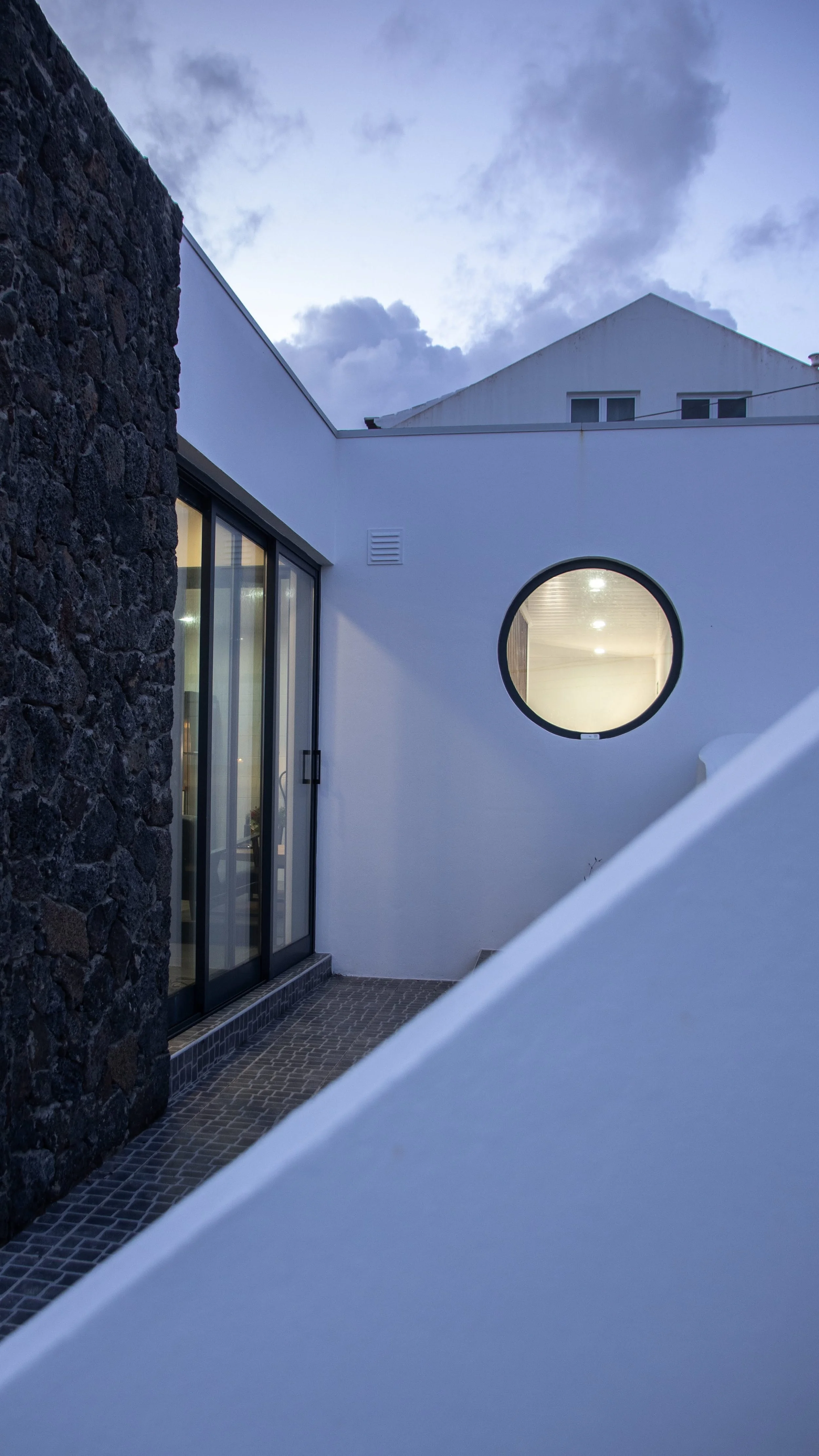 A modern building exterior with a large circular window, sliding glass door, stone wall, and cobblestone ground, with the sky and clouds in the background.