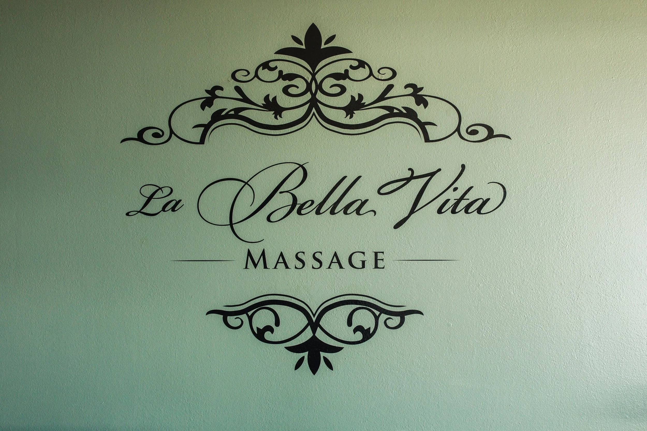 Decorative sign on a wall reading "La Bella Vita Massage" with ornate black design elements on a light green background.