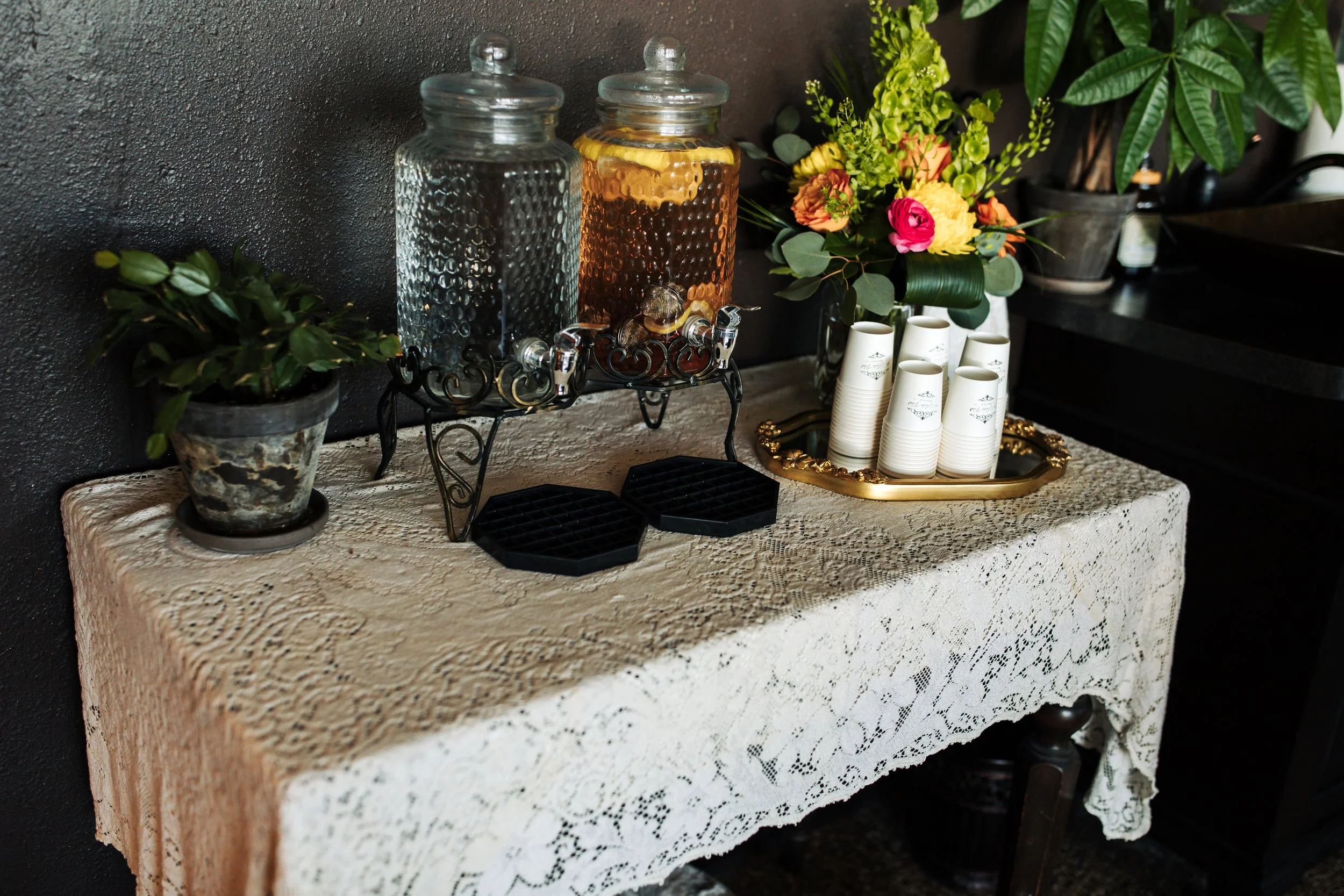 A lace-covered table with a floral arrangement, glass beverage dispensers, and ceramic cups.
