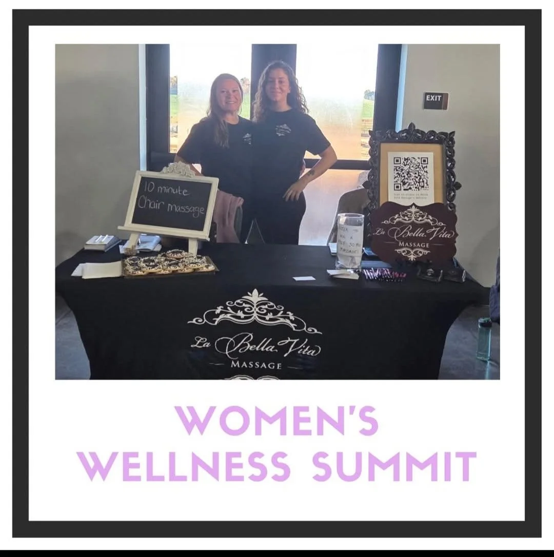Photograph of two women behind a table at a Women's Wellness Summit. The table has a sign advertising a 10-minute chair massage and features decorations and business cards. The women are smiling, standing inside a venue with large windows showing an outdoor landscape.