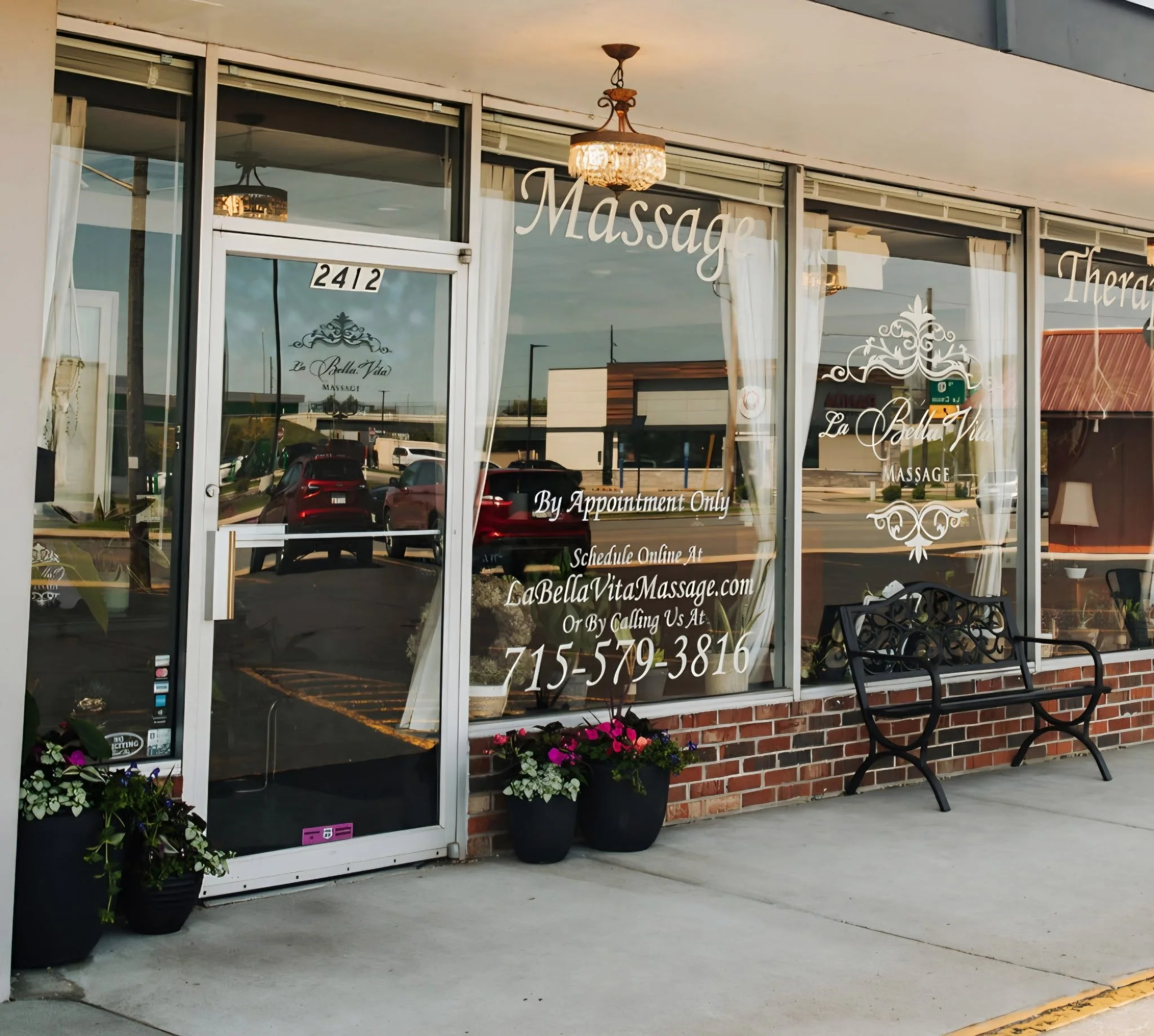 The exterior of a massage therapy clinic with large glass windows and white curtains. The door has a sign with the address 2412, and the windows display the business name 'La Bella Vita Massage.' There are flower pots with colorful flowers outside, and a black metal bench is placed on the sidewalk in front of the windows.