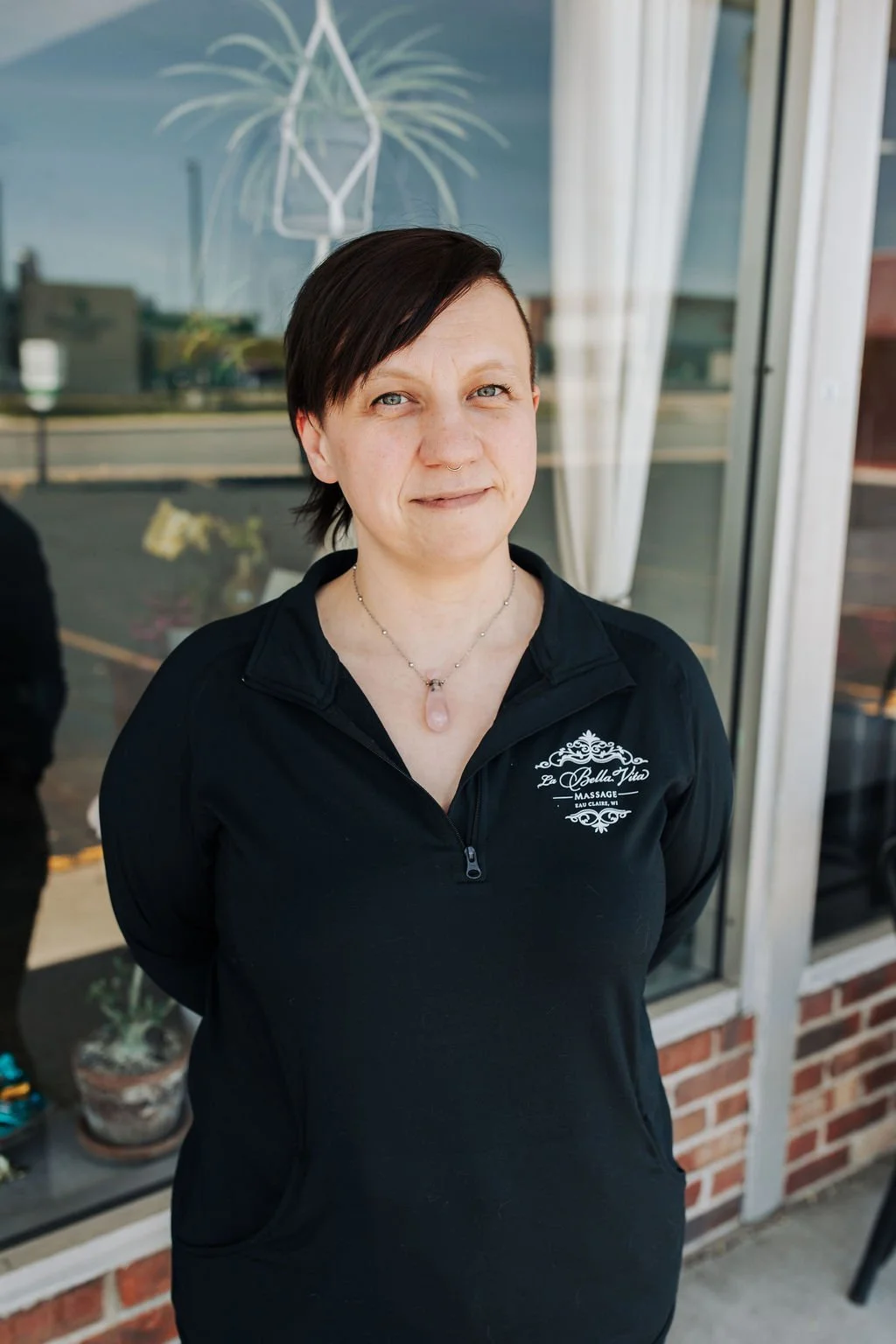 A woman with short dark hair standing outside a building, wearing a black zip-up jacket with a logo that reads "La Bella Vita Massage". She is also wearing a necklace with a pink stone pendant.
