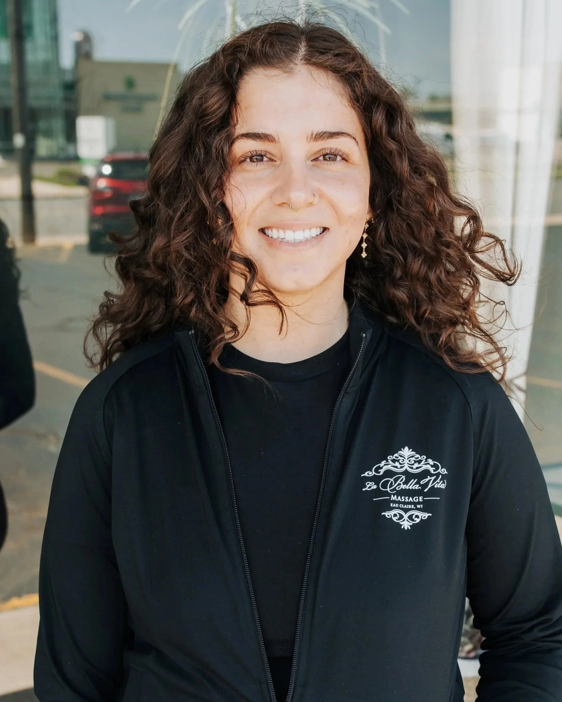A woman with curly brown hair smiling in front of a glass window. She is wearing a black zip-up jacket with a logo that reads 'La Bella Vita Massage Eau Claire, WI' on the chest.