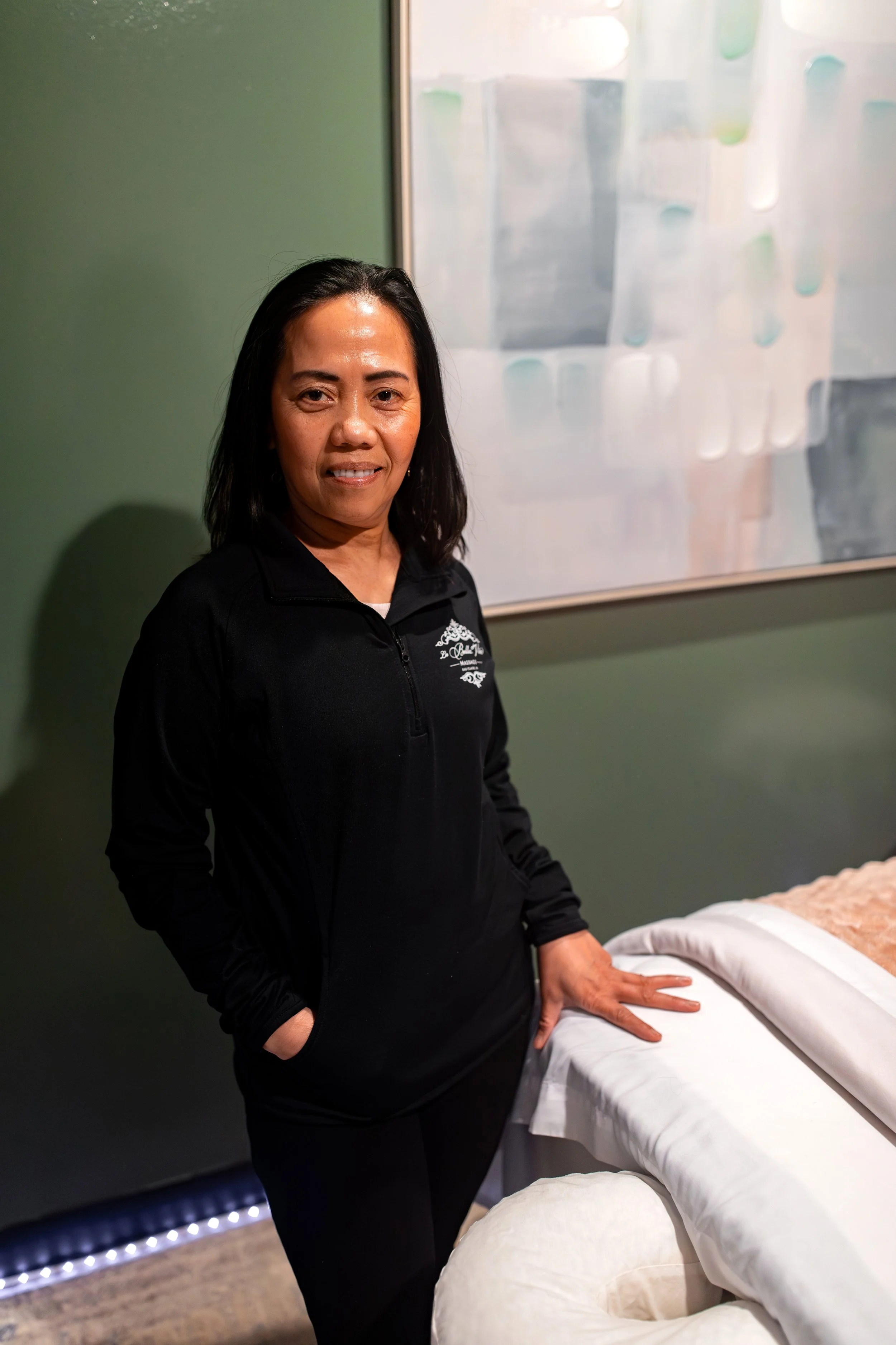 A woman standing next to a massage table in a wellness or massage center, smiling at the camera.