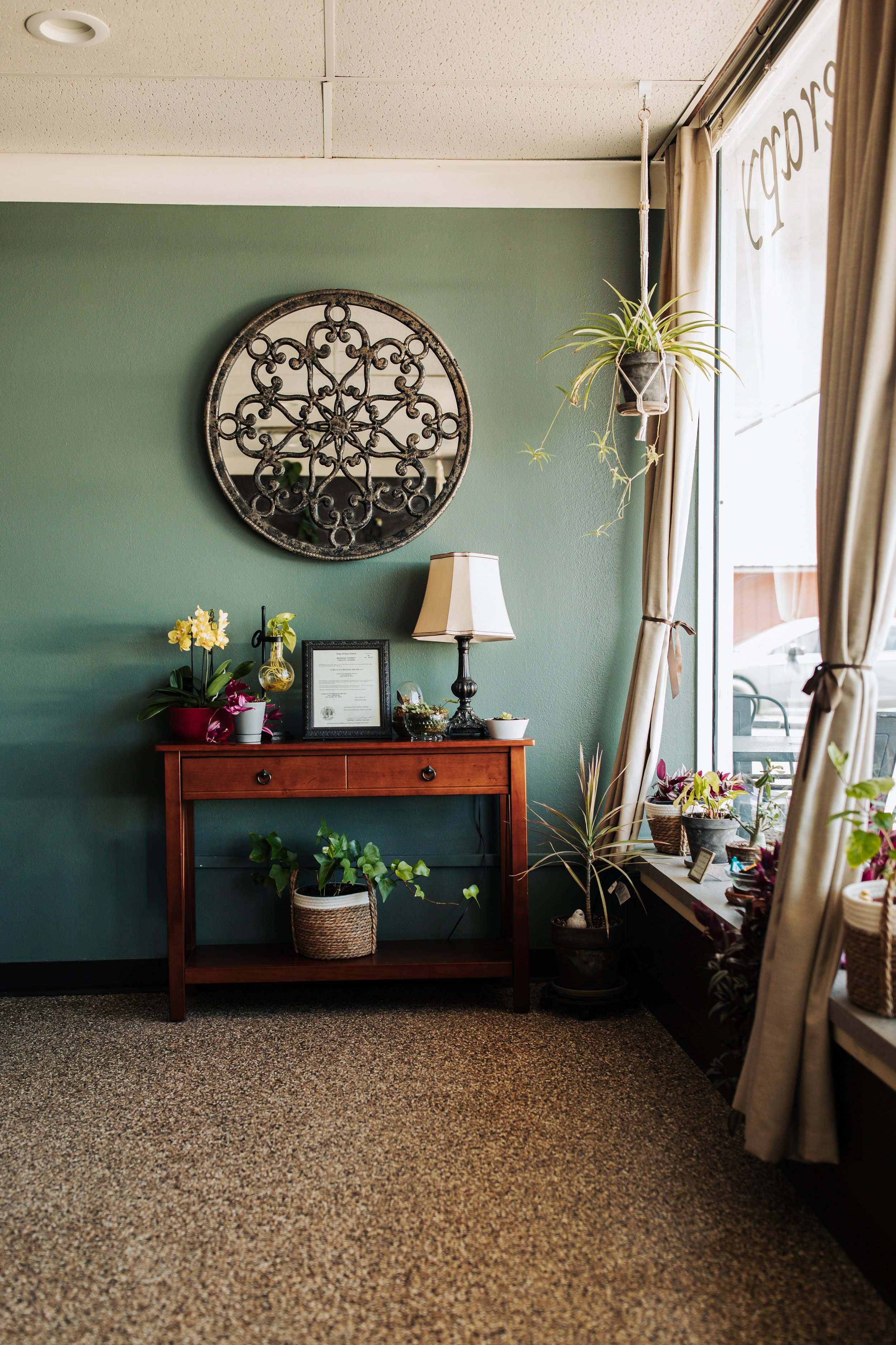 Interior of a room with a green wall, a wooden table with plants and a framed document, a lamp, and a hanging potted plant near large windows with curtains, carpeted floor.