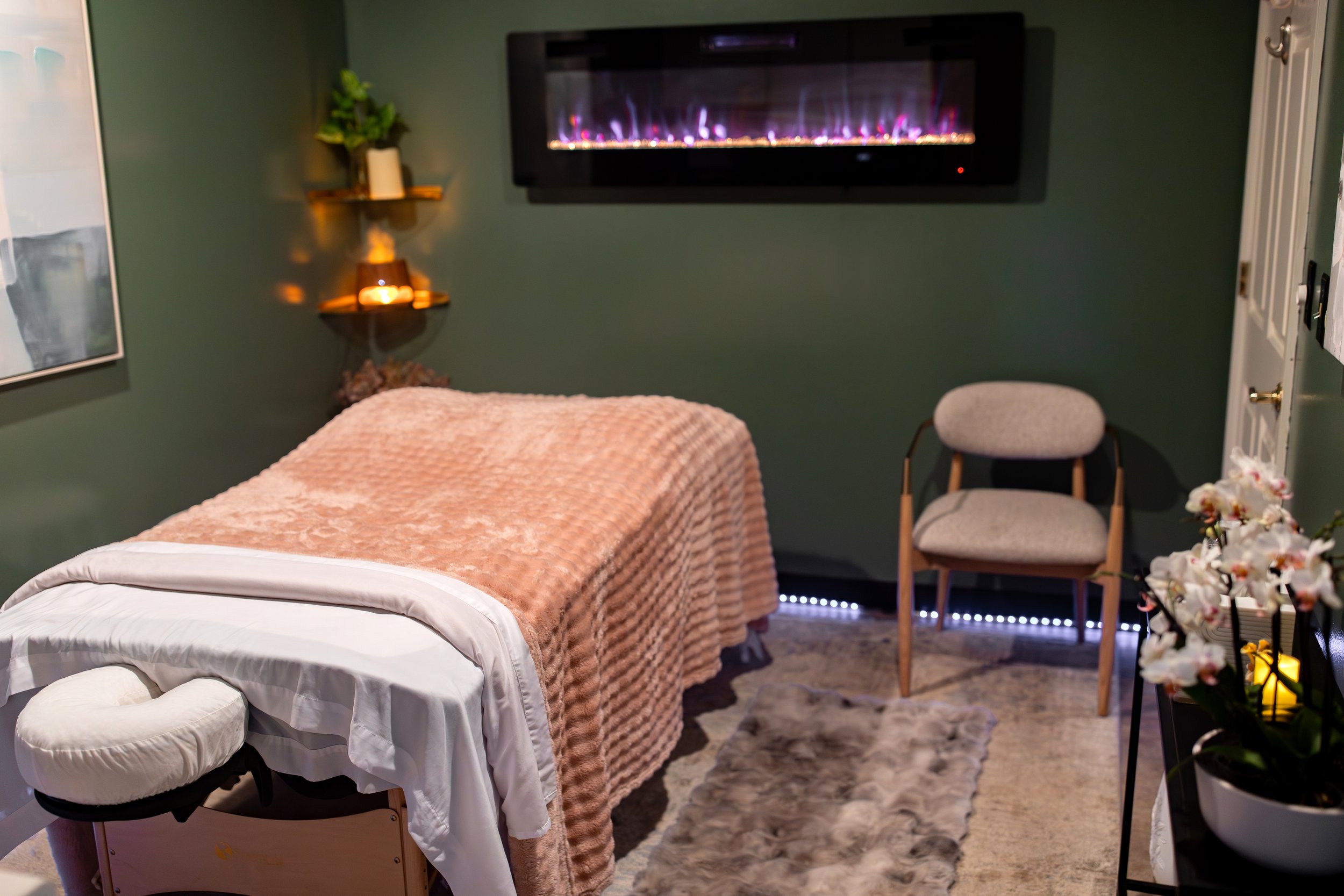 A cozy bedroom with a massage table covered in a blush pink blanket, a wall-mounted electric fireplace, a chair, and decorative plants and candles.