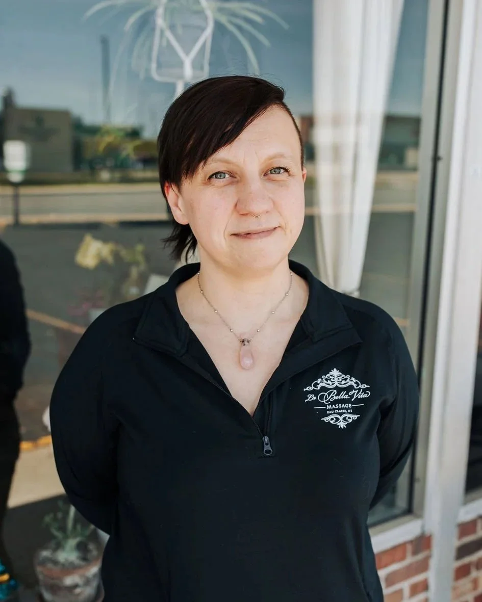A woman standing outside in front of a glass door with white curtains, wearing a black zip-up jacket with a logo and the words "La Bella Vita Massage.” She has short dark hair with an asymmetrical cut and a pink gemstone necklace.