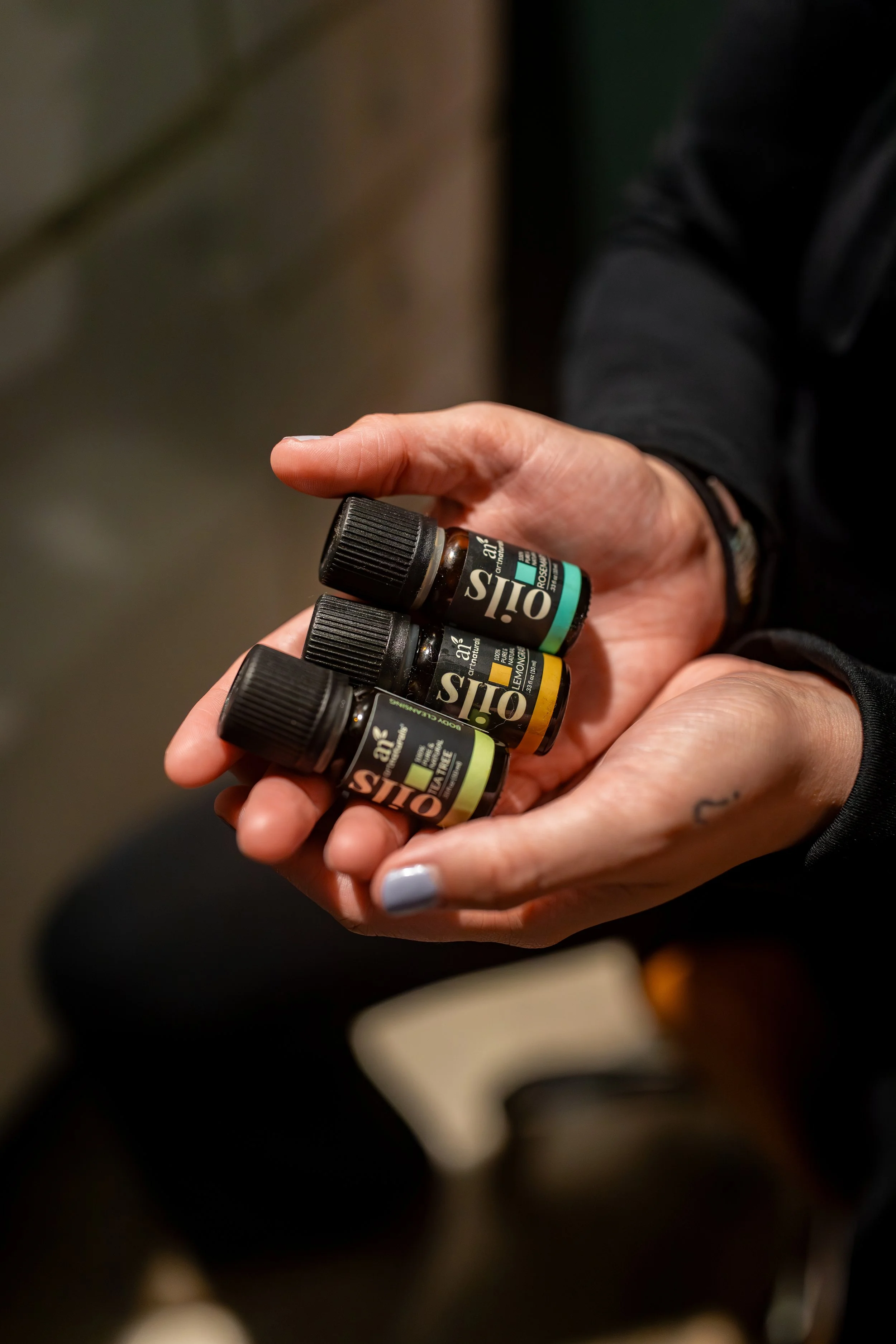 Close-up of a person's hand holding several small essential oil bottles with black caps and labels showing brand and scent names.