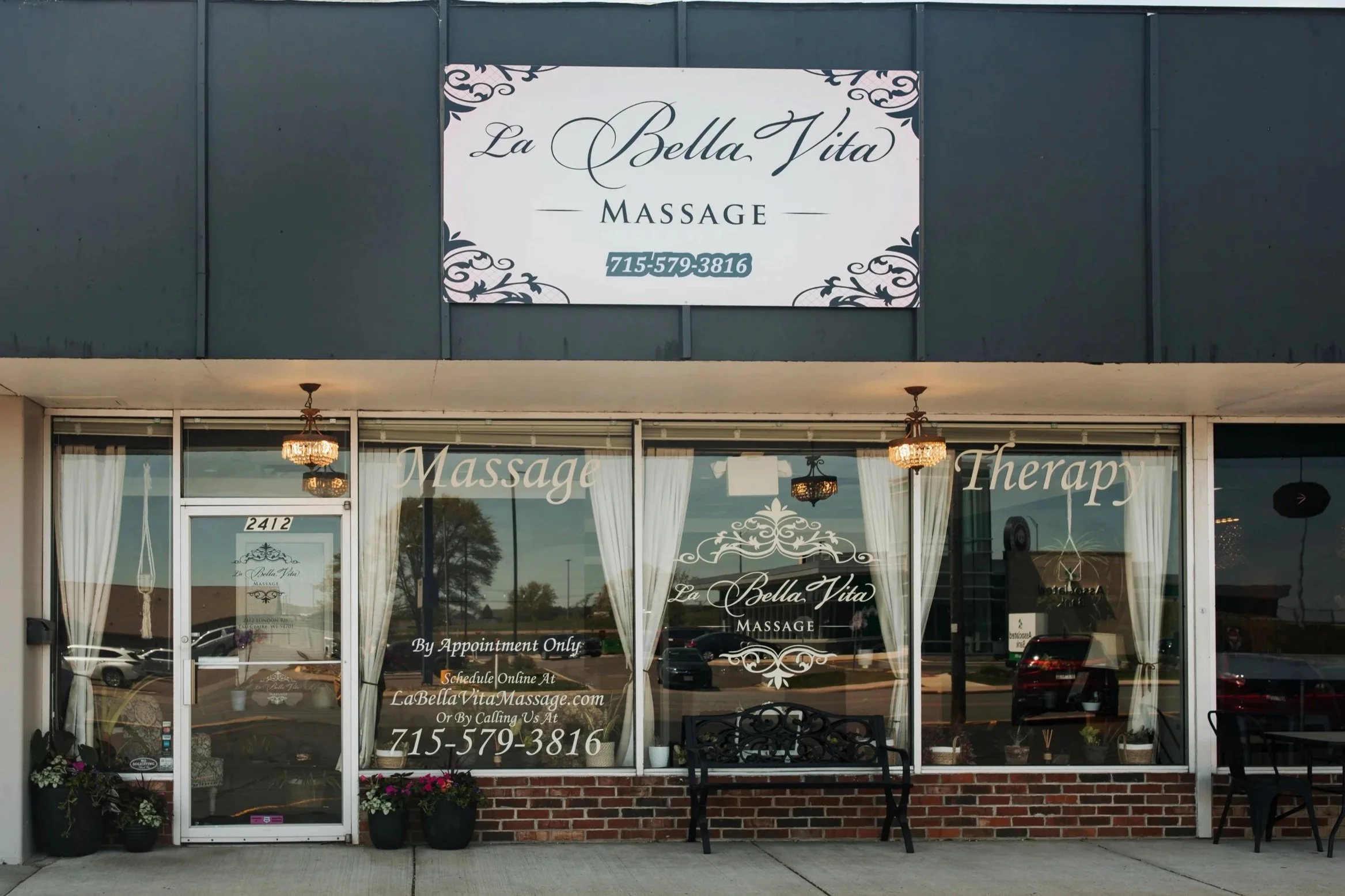 Front view of La Bella Vita Massage spa with a sign board on the upper section displaying the name and phone number, glass windows with curtains, small outdoor seating area with a bench, potted plants, and a chandelier inside.