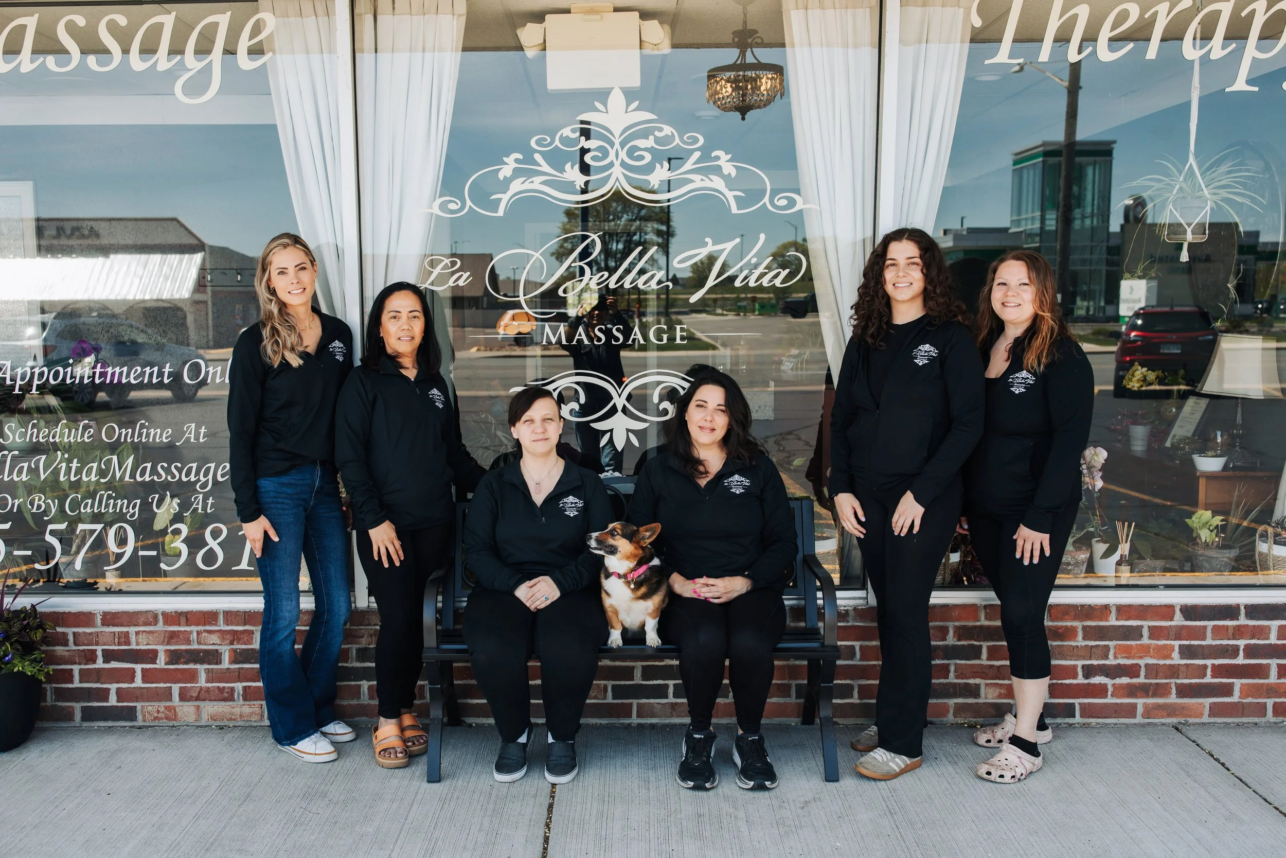 Group of five women and one dog standing and sitting in front of a massage spa window with white curtains, a chandelier, and a plant, outside a brick building.