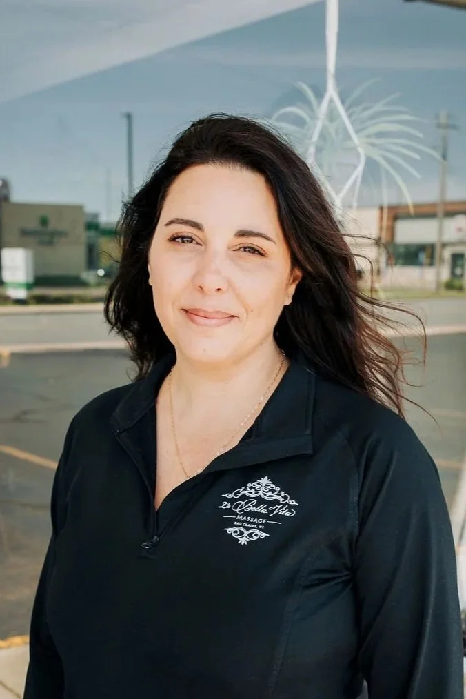 A woman with dark hair standing outside wearing a black zip-up shirt with a logo for Bella Vita Massage.