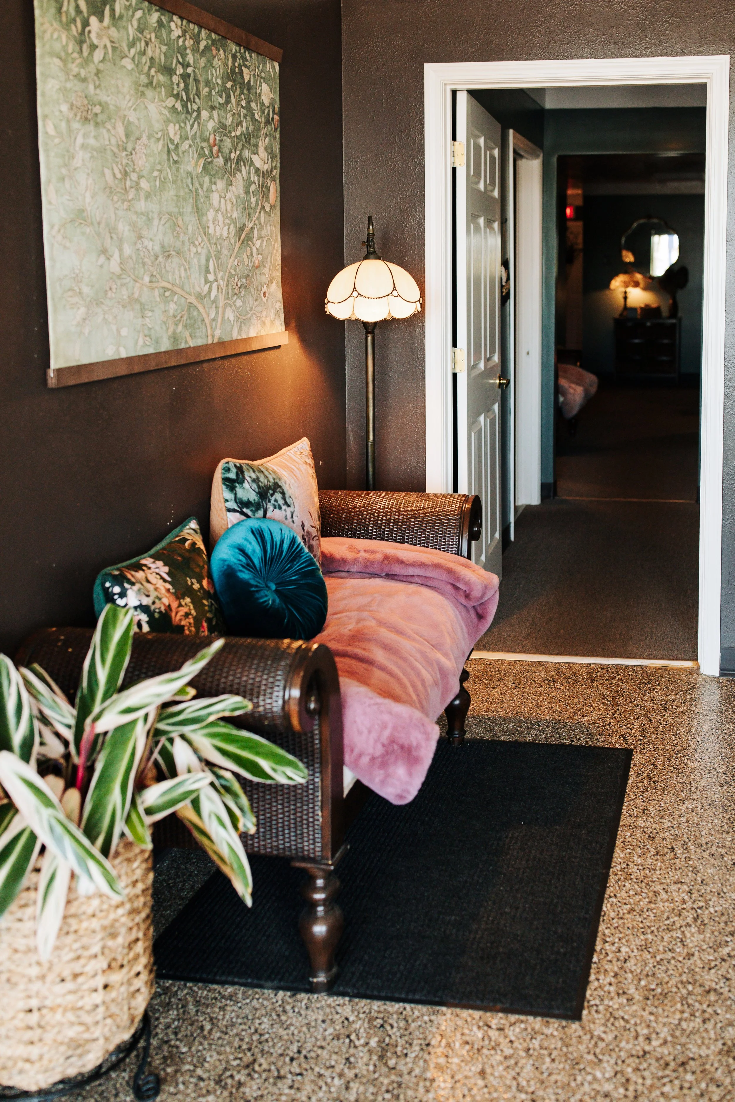 A cozy living room corner with a pink velvet couch decorated with various colorful pillows, a tall vintage floor lamp with a scalloped shade, a large leafy plant in a woven basket, and a dark wall with a floral tapestry hanging on it. An open doorway reveals a dimly lit room with a dresser and other furniture.