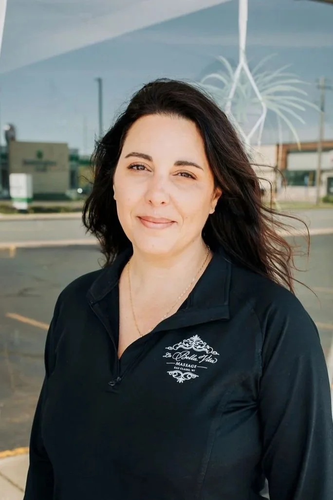 A woman with dark, wavy hair stands outside, wearing a black zip-up jacket with a logo that says 'La Celle Vida Massage'. She is smiling gently at the camera, and the background shows a parking lot and some building reflections on the glass window behind her.