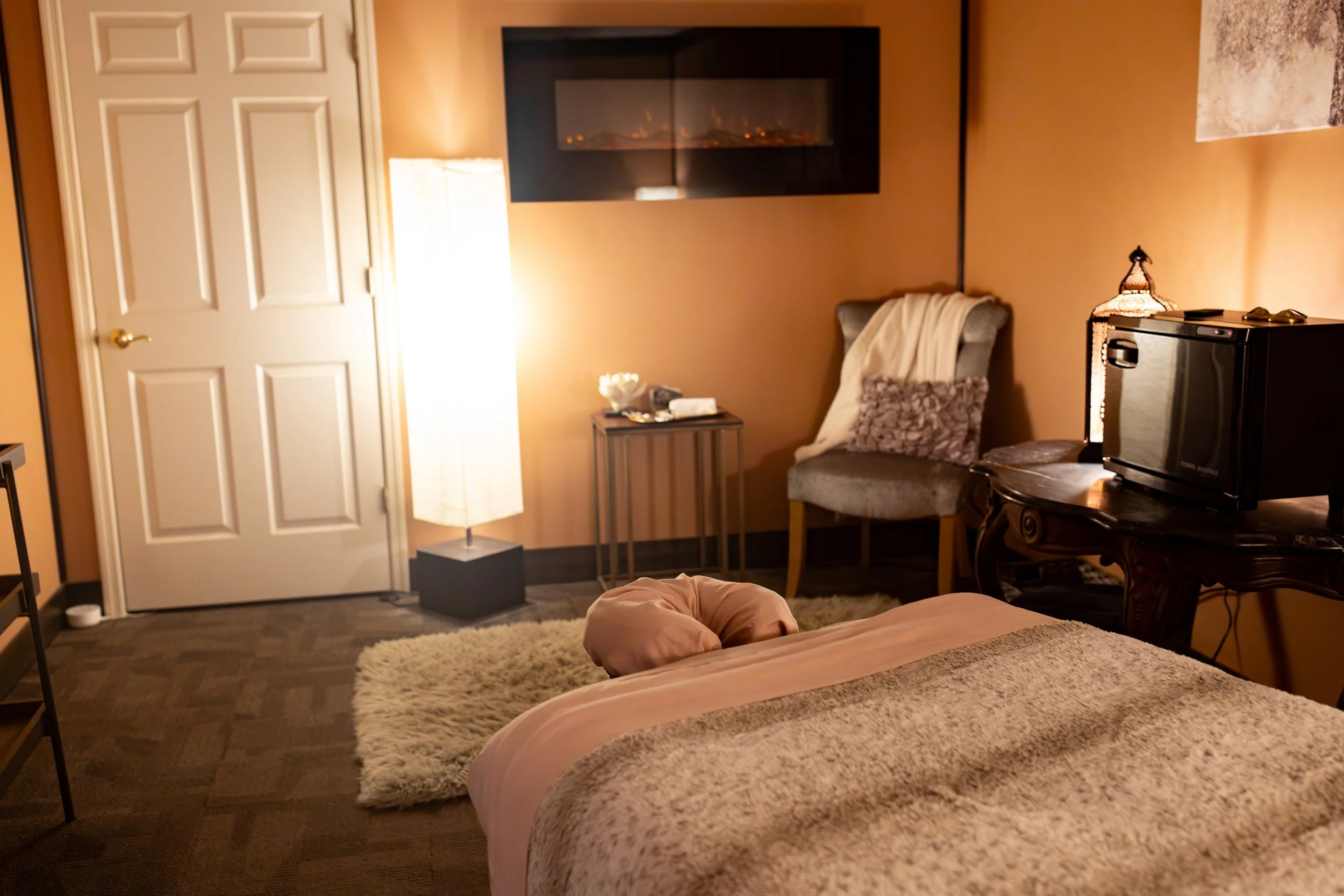 A cozy bedroom with a bed covered in soft beige and pink bedding, a plush rug on the dark wood floor, a side table with a lamp and decorative items, a chair with a blanket and pillows, a modern wall-mounted fireplace, a small refrigerator, and a floor lamp, creating a warm, inviting atmosphere.