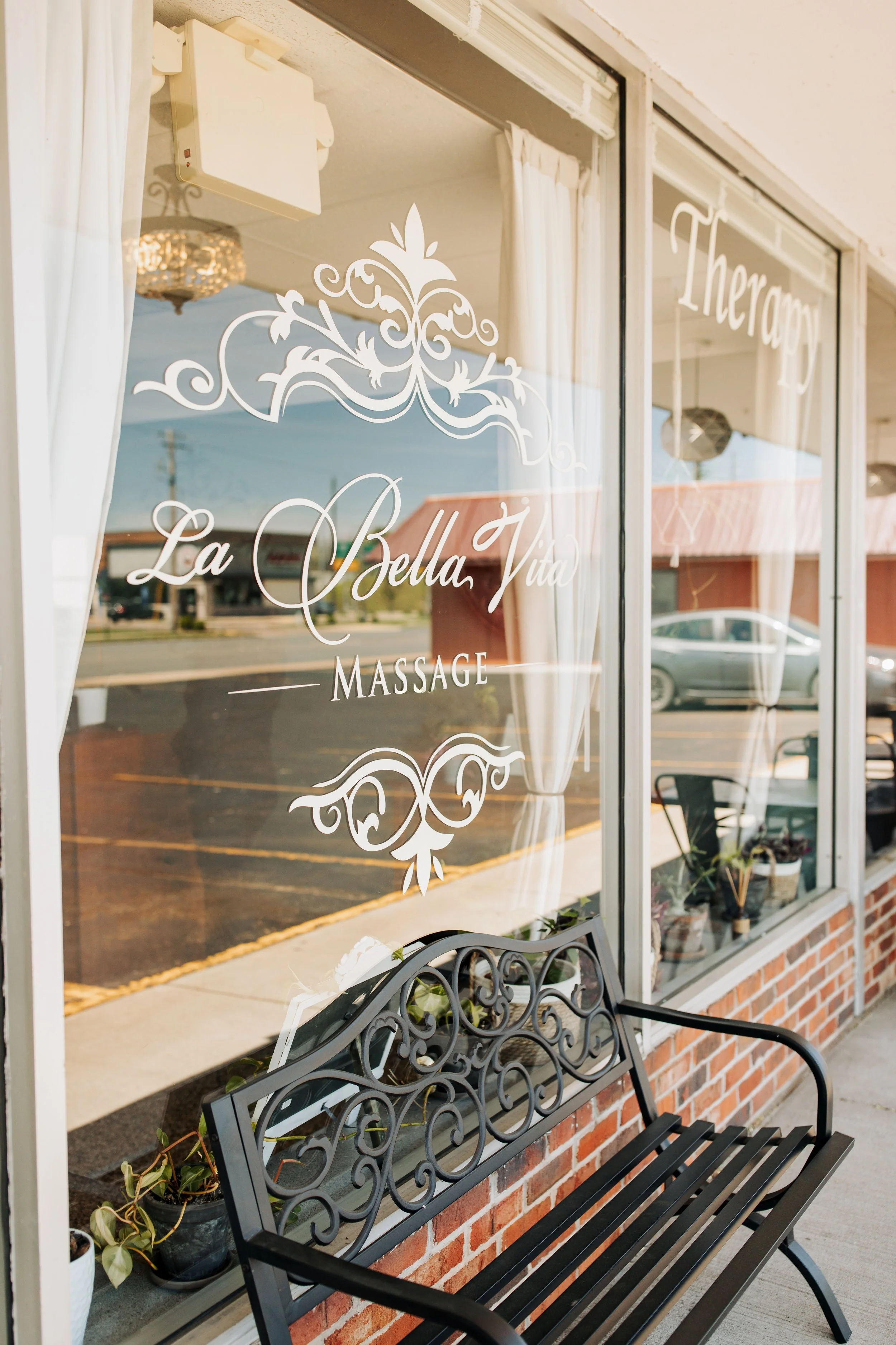 The image shows the front window of a massage business called 'La Bella Vita Massage'. The window has white decorative lettering and a floral design. There is a black iron bench with a plant potted in black beside it outside the window.