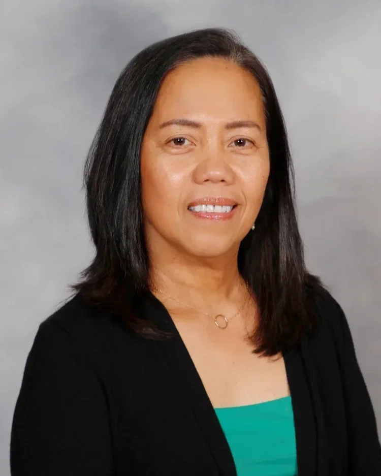 A woman with shoulder-length black hair, wearing a black blazer over a green top, and a gold necklace, smiling against a gray background.