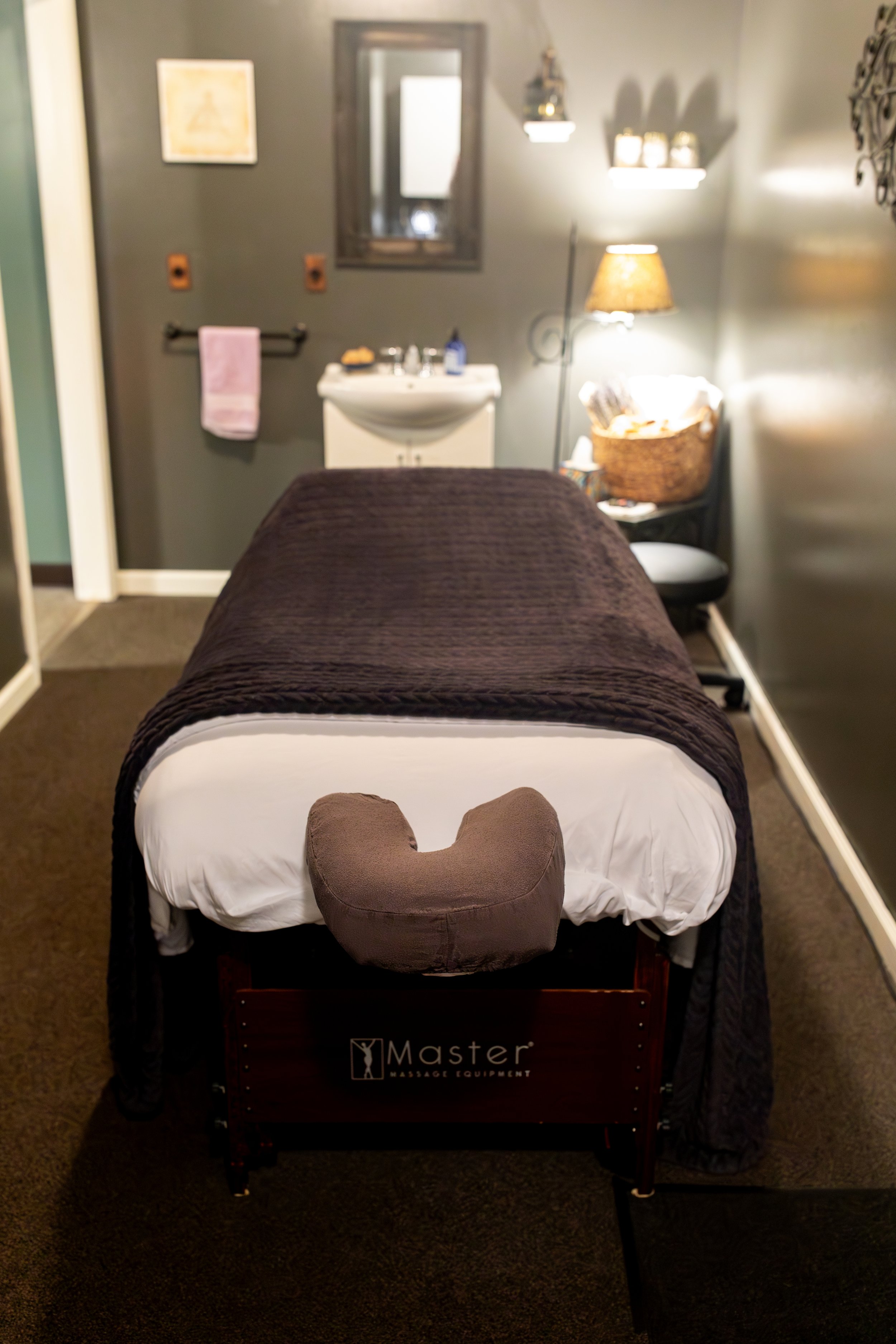 A massage table covered with a dark brown blanket, with a brown heart-shaped pillow at the head, in a room with soft lighting, wall decorations, and a massage equipment cart labeled 'Master Massage Equipment'.