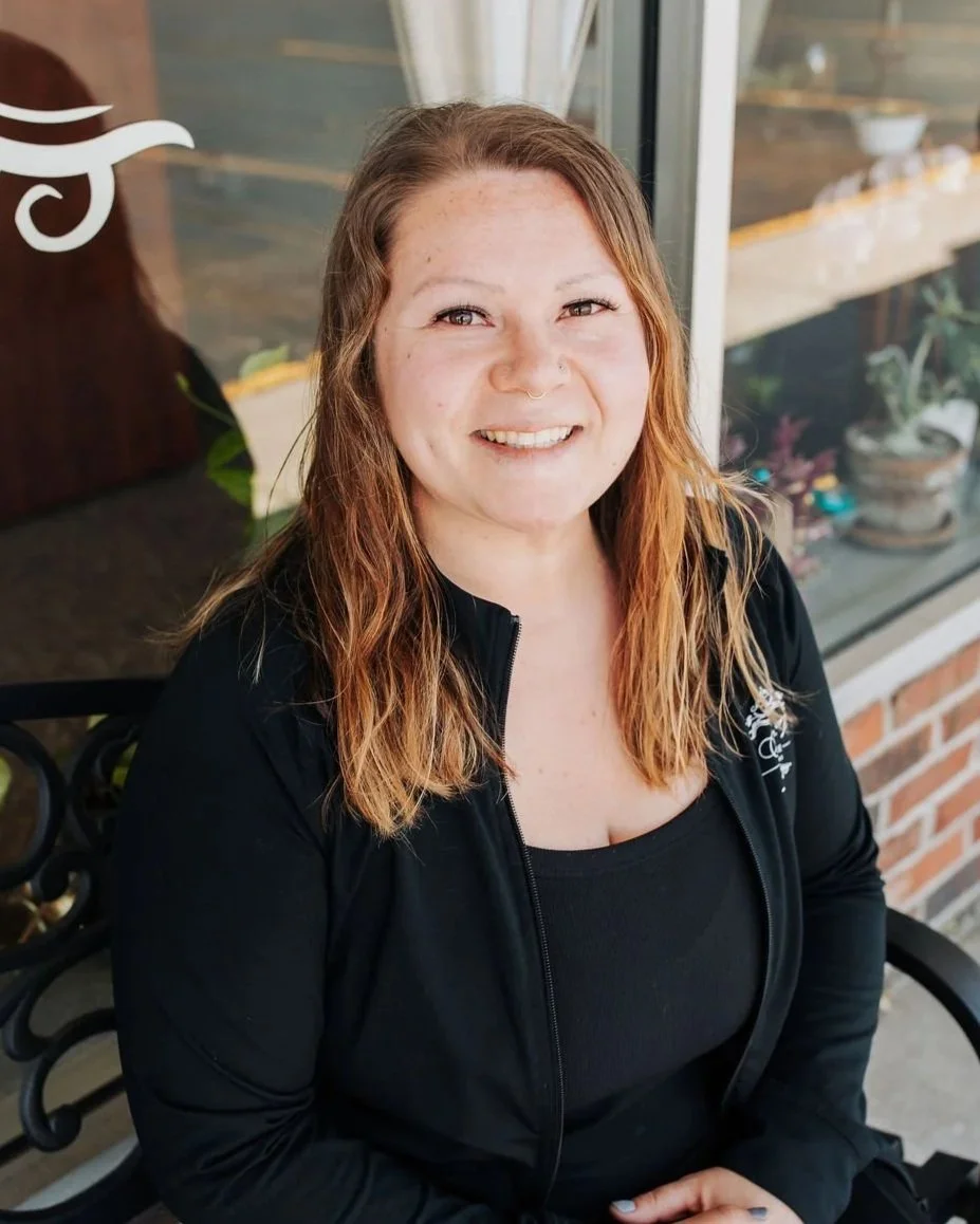 A woman with long, wavy, auburn hair and fair skin, smiling while sitting outdoors on a black wrought iron bench, with a brick wall and window behind her.