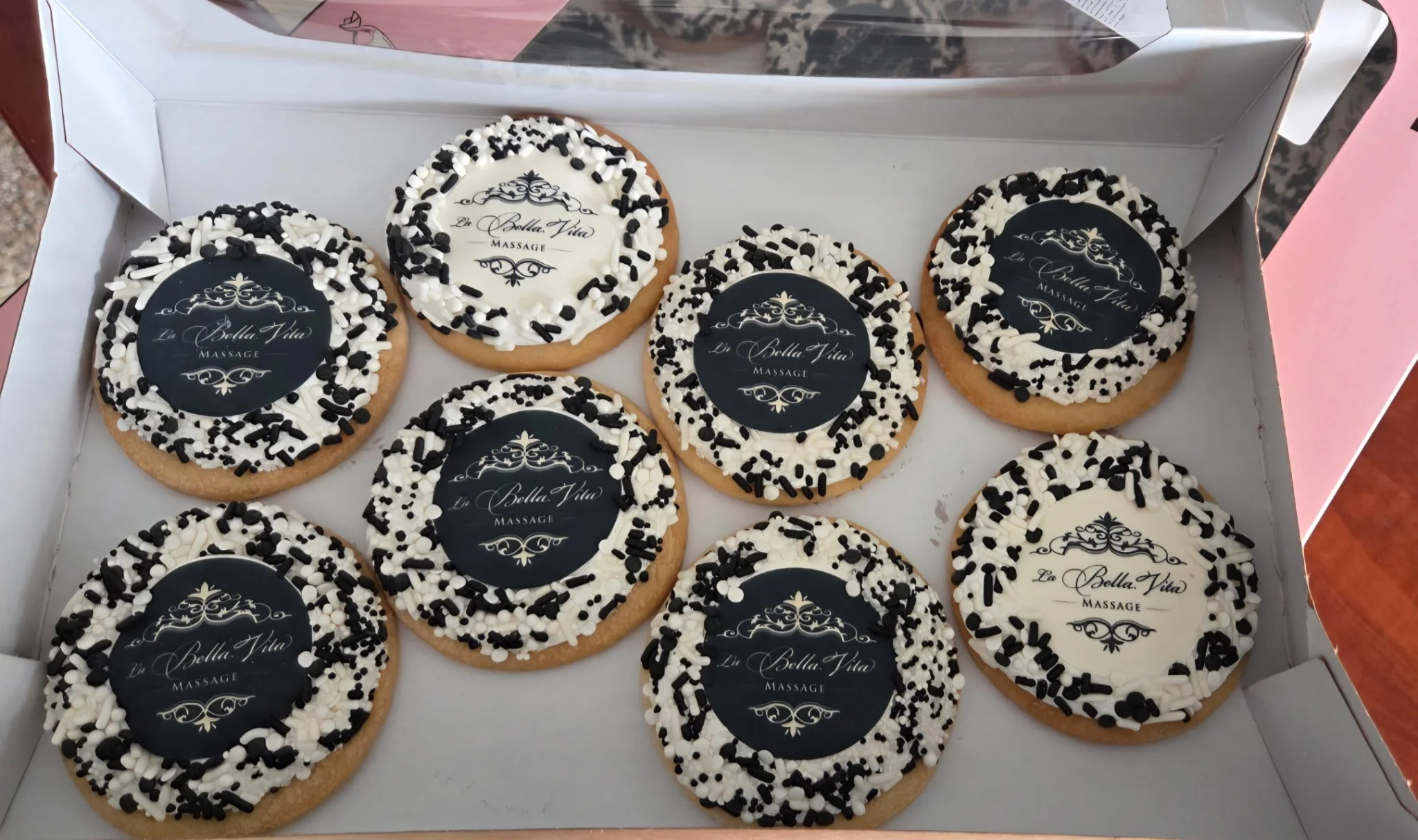 A box of cookies decorated with black and white sprinkles and a printed edible logo for 'La Bella Vita Massage'.
