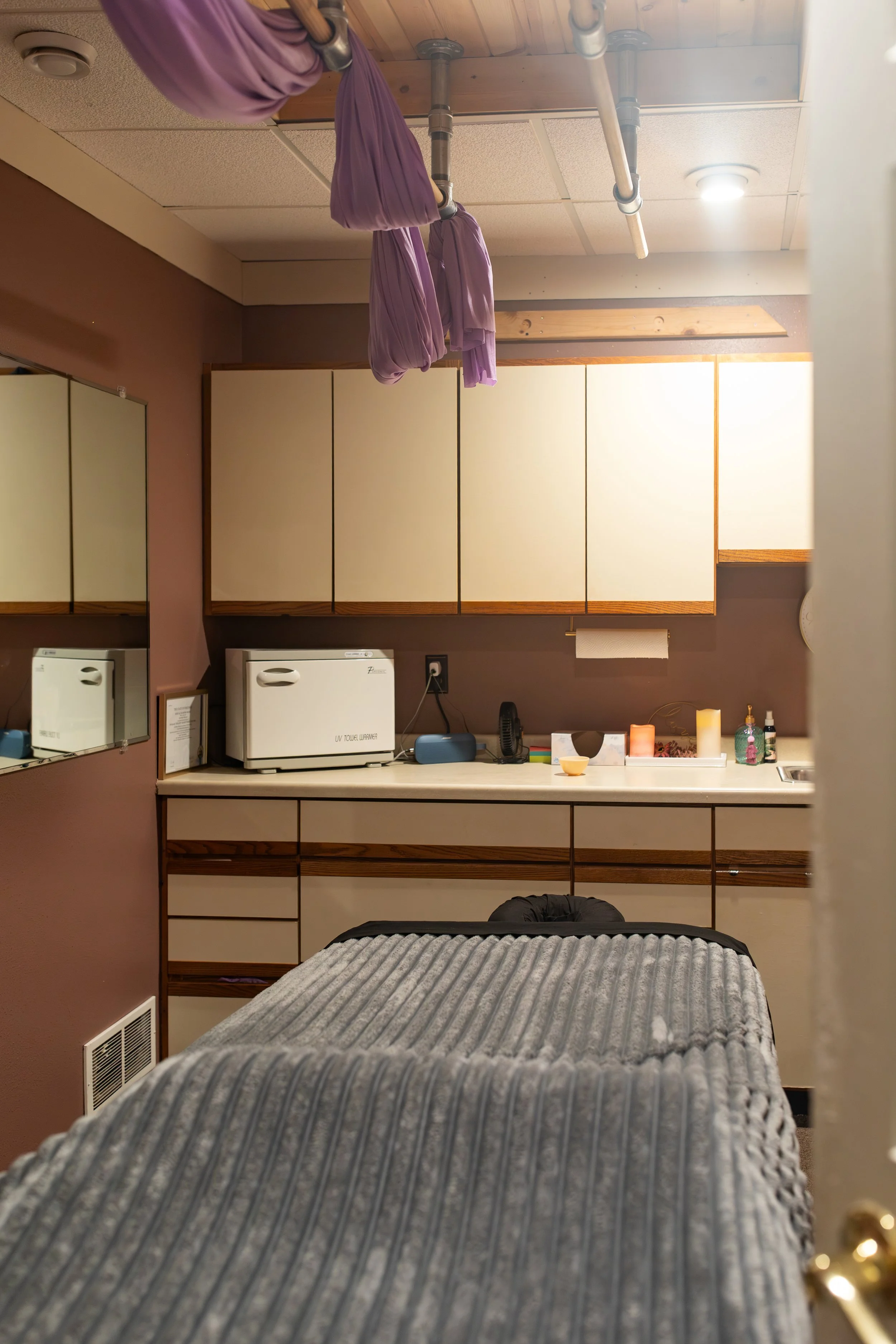 A bedroom with a bed in the foreground covered with a gray knitted blanket, a desk area with cabinets, a microwave, a phone, candles, and decorative items, and purple fabric hanging from pipes attached to a wooden ceiling.