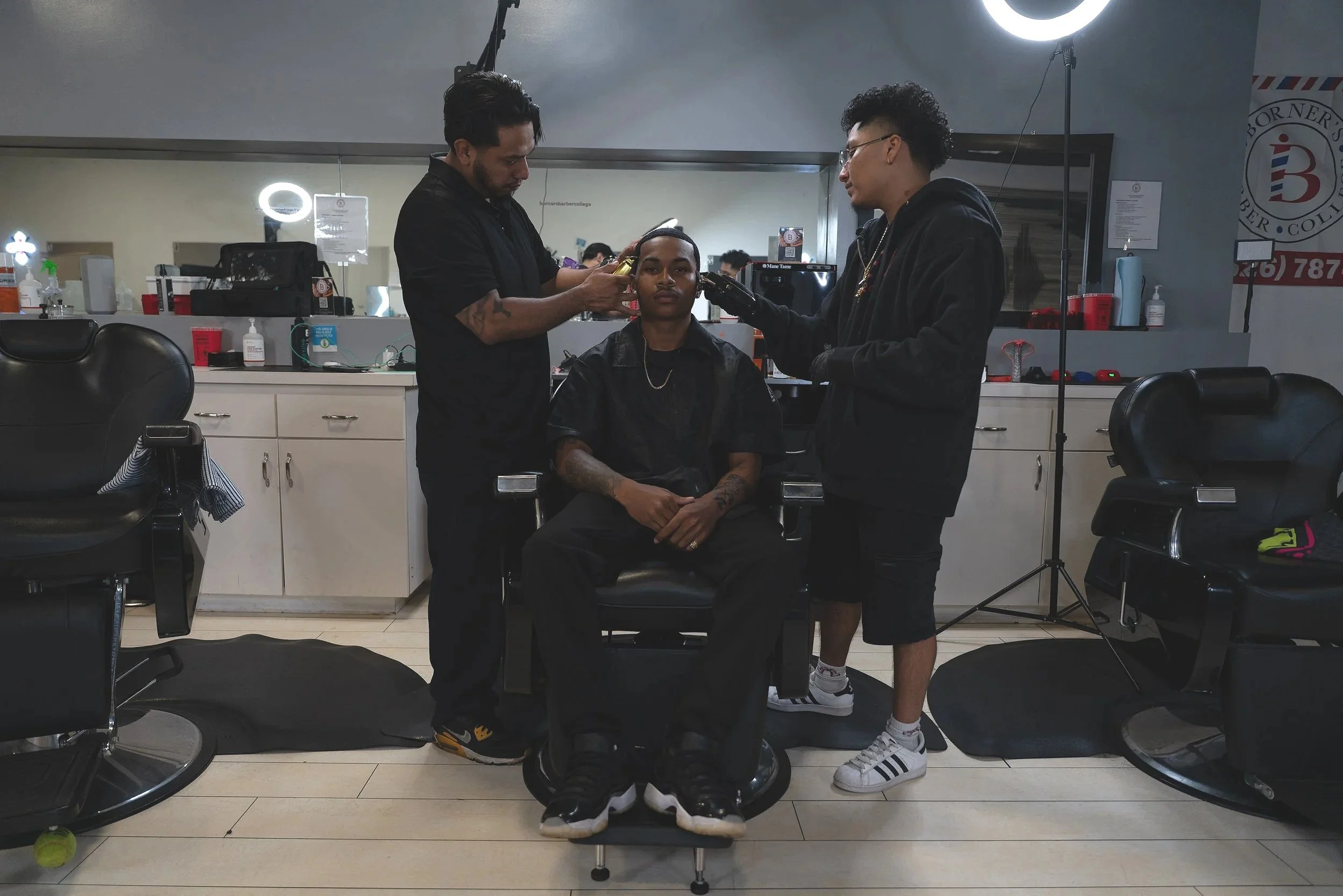 A man sitting in a barber chair at a barbershop, getting a haircut from two barbers. The barber on the left is using a hair clipper, and the barber on the right is holding a spray bottle. The barbershop has black chairs, a mirror, and various barber tools and products on the counter behind them.