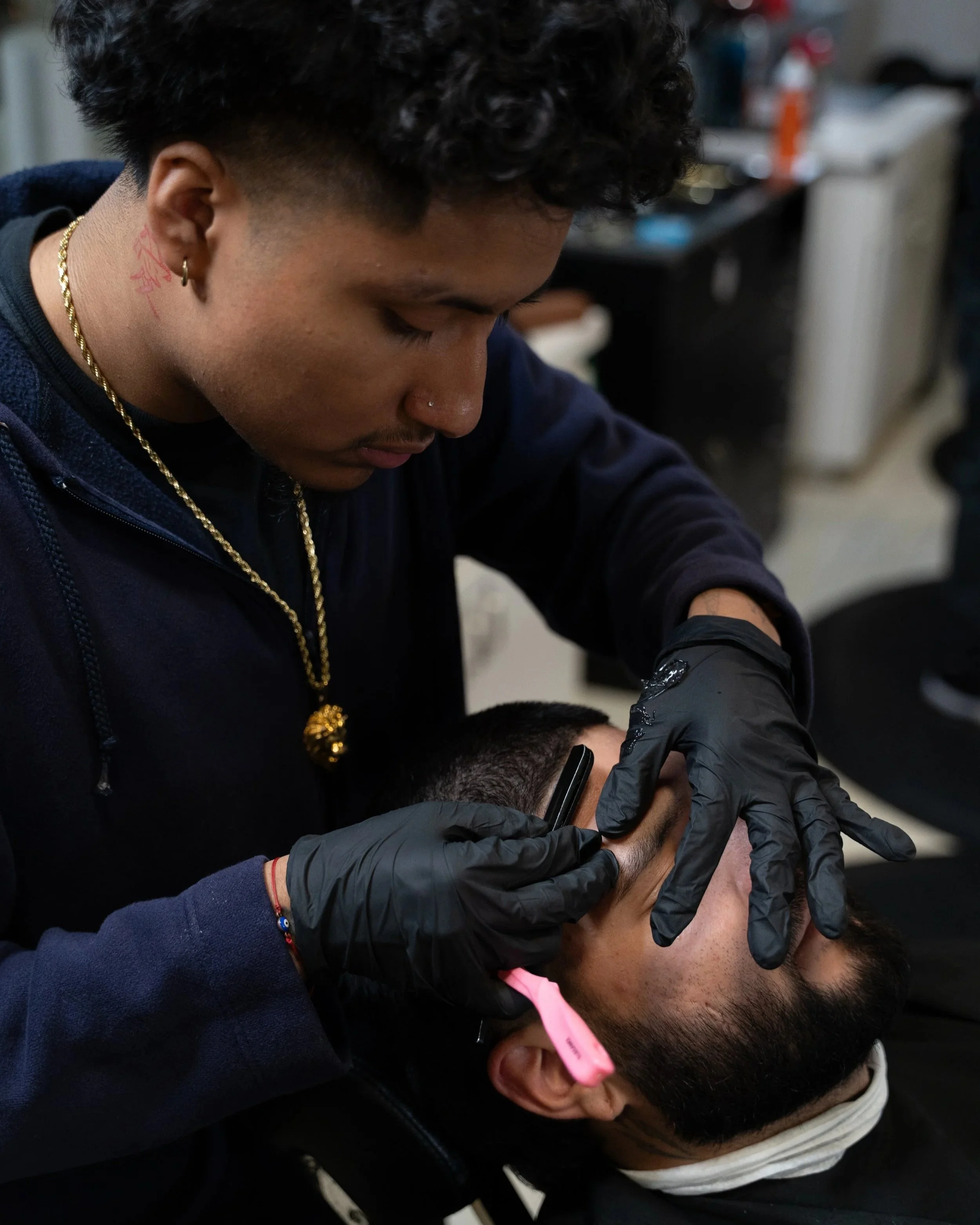 Close-up of a barber tattoo, earrings, gold necklace, and a black glove, working on giving a haircut to a young man with glasses and a pink barber cape, in a barbershop.