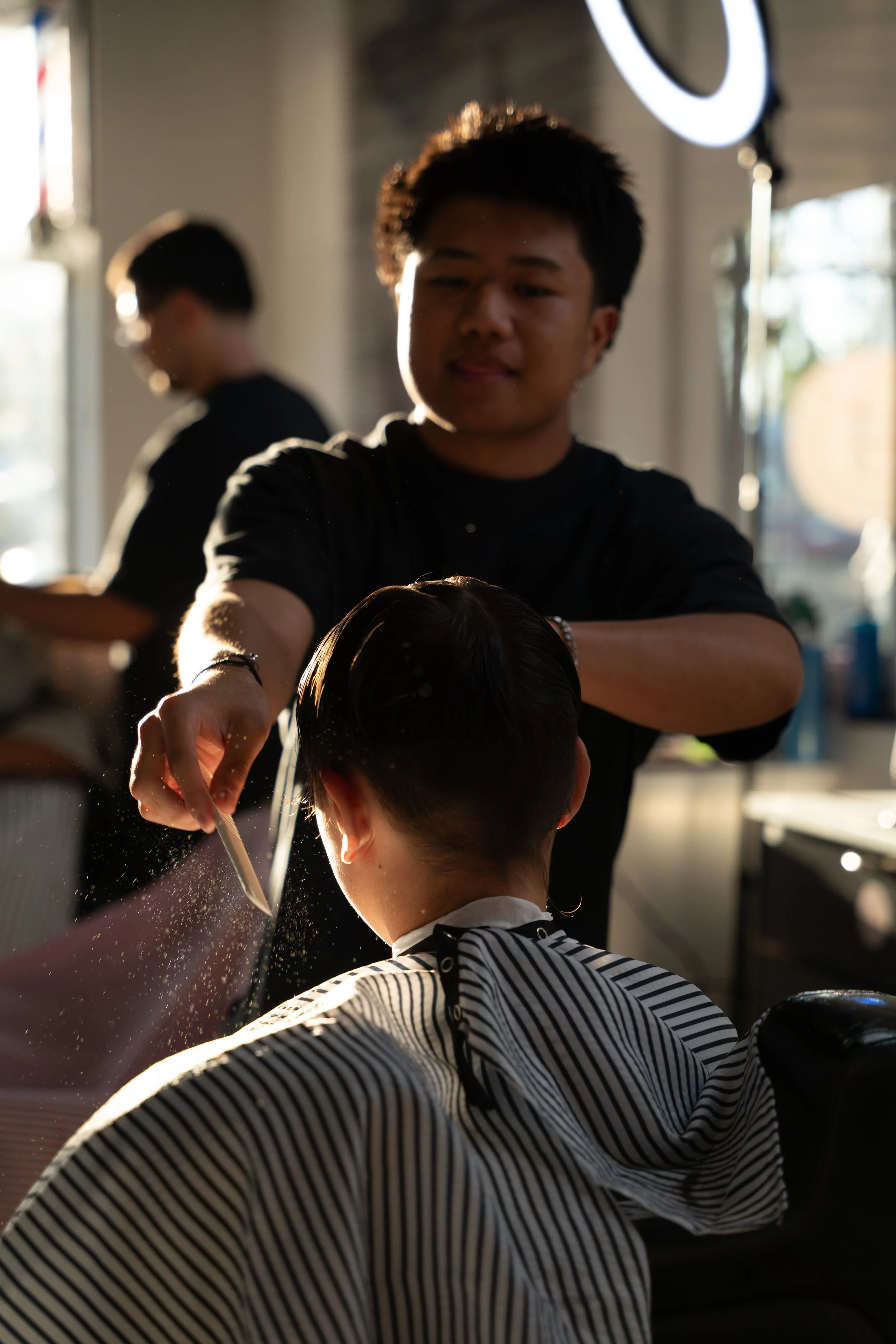 A hairstylist applying hair treatment to a client's hair in a salon. The salon is illuminated by warm backlighting, with a mirror and ring light visible in the background.