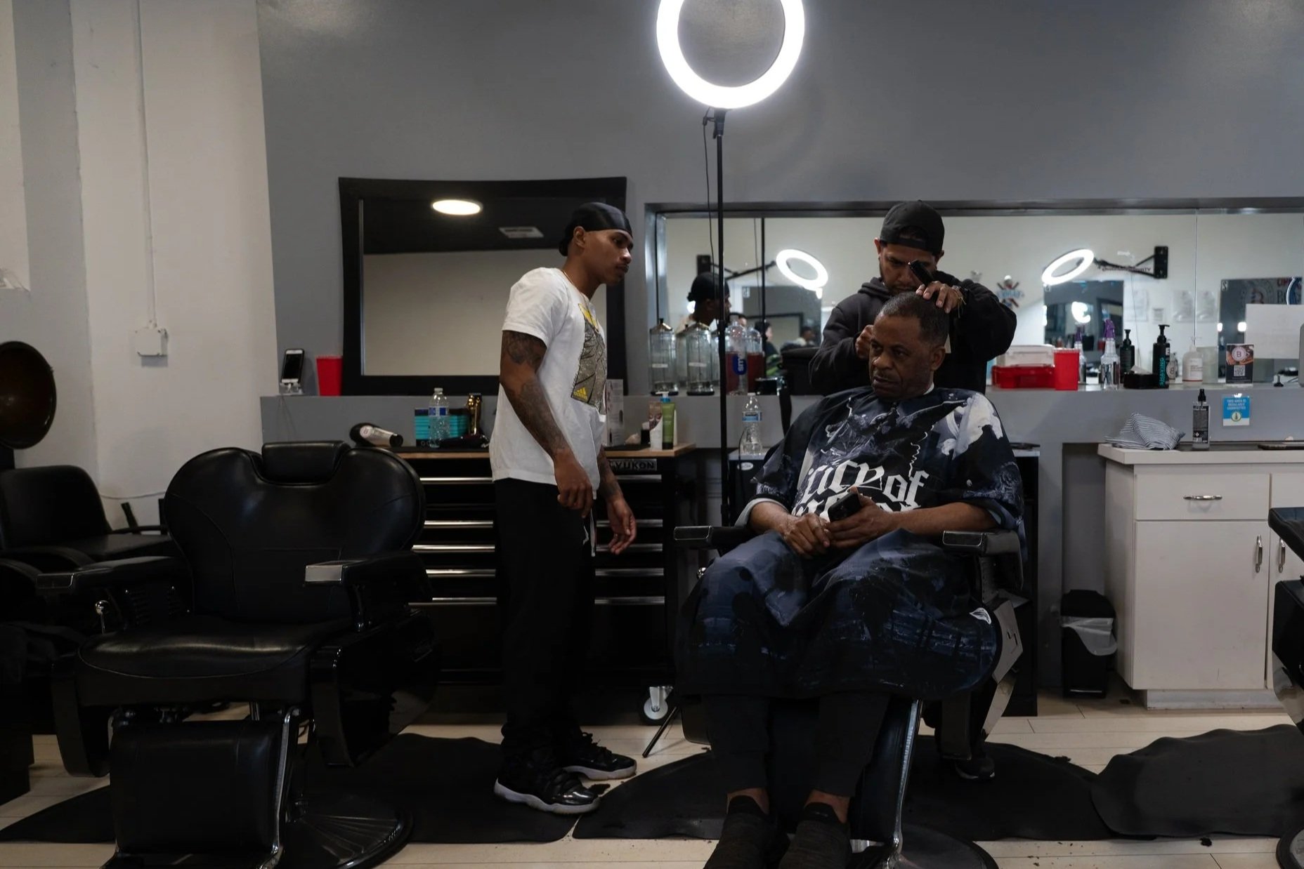 A man is sitting in a barber chair at a barbershop, looking at his phone, while a barber is styling his hair. Another man stands nearby, observing. The barbershop has black chairs, a mirror, and various hair products on the counter.