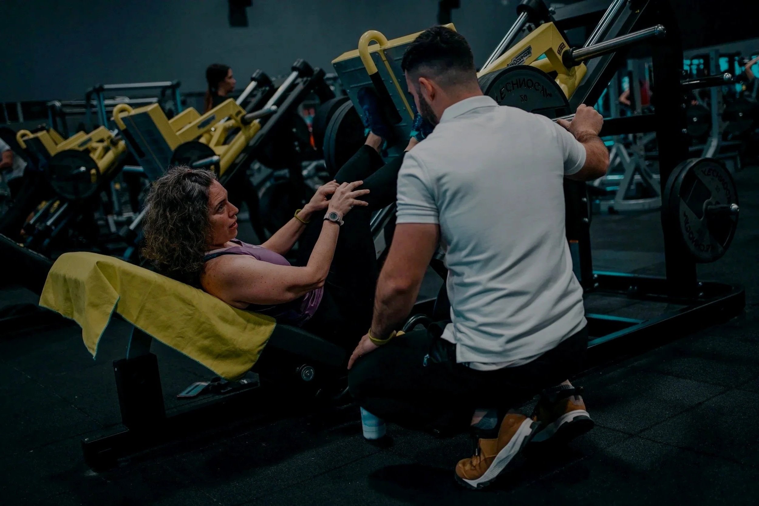A woman lying on a workout bench receiving instruction from her fitness coach (Riley May). The scene includes gym equipment such as weight plates and other fitness machines, with a few people visible in the background.