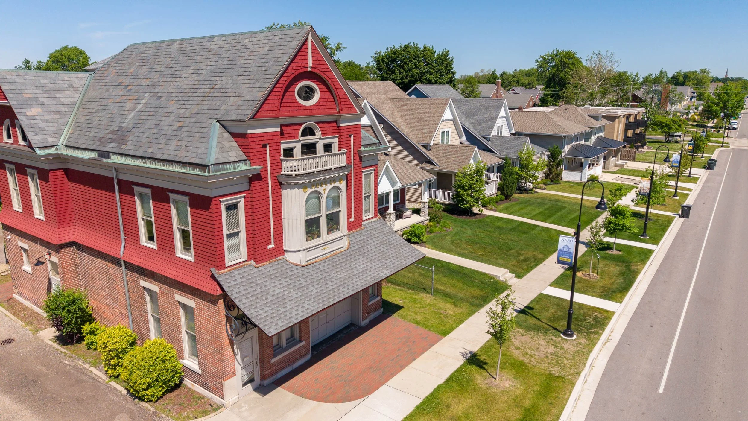 Aerial view of a red Victorian-style house with a gabled roof and multiple windows, neighboring houses with similar architectural styles, a sidewalk, street lamps, and a landscaped grassy area in a suburban neighborhood on a sunny day.
