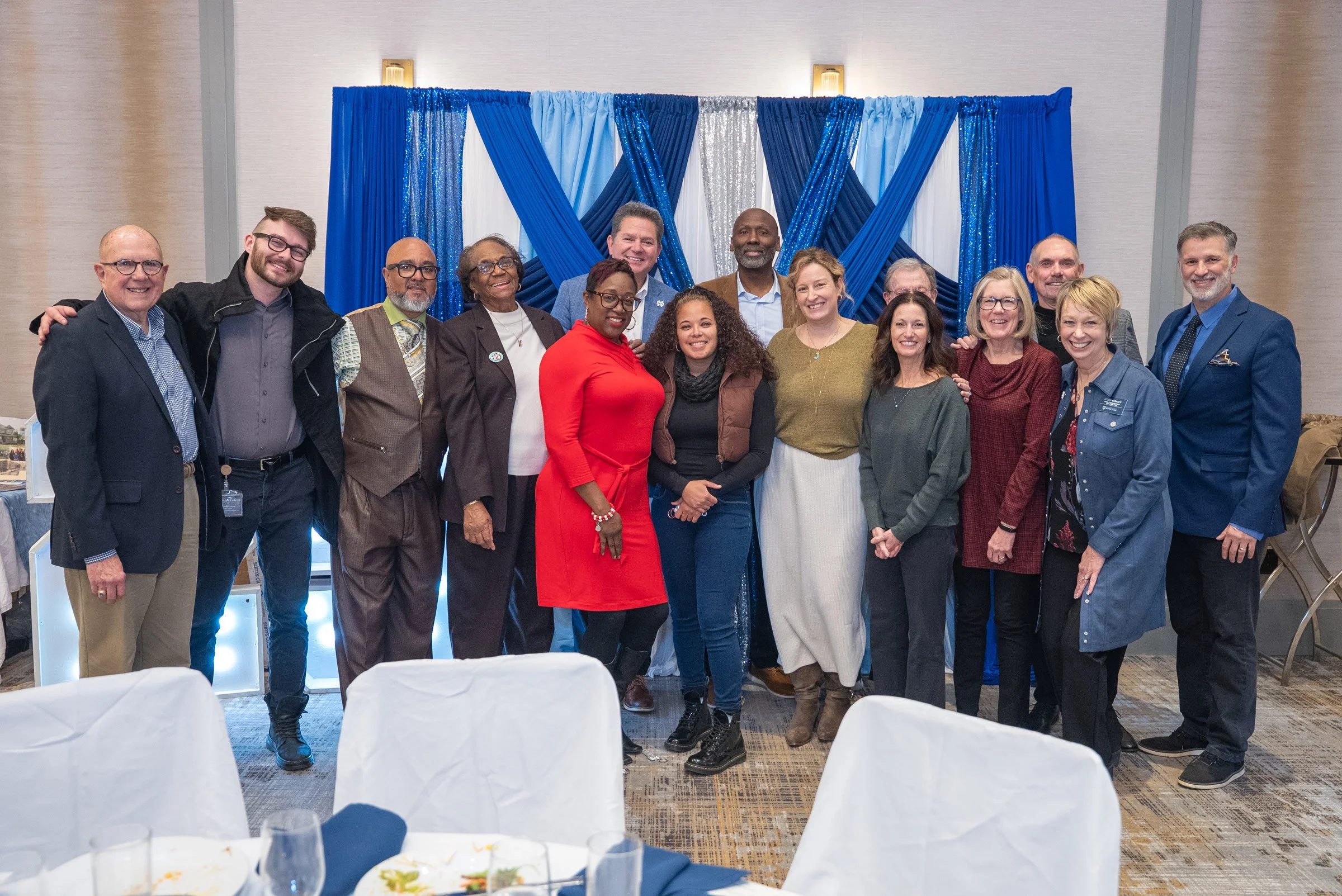 Group of diverse people standing together at an indoor event with a blue and silver decorated backdrop.