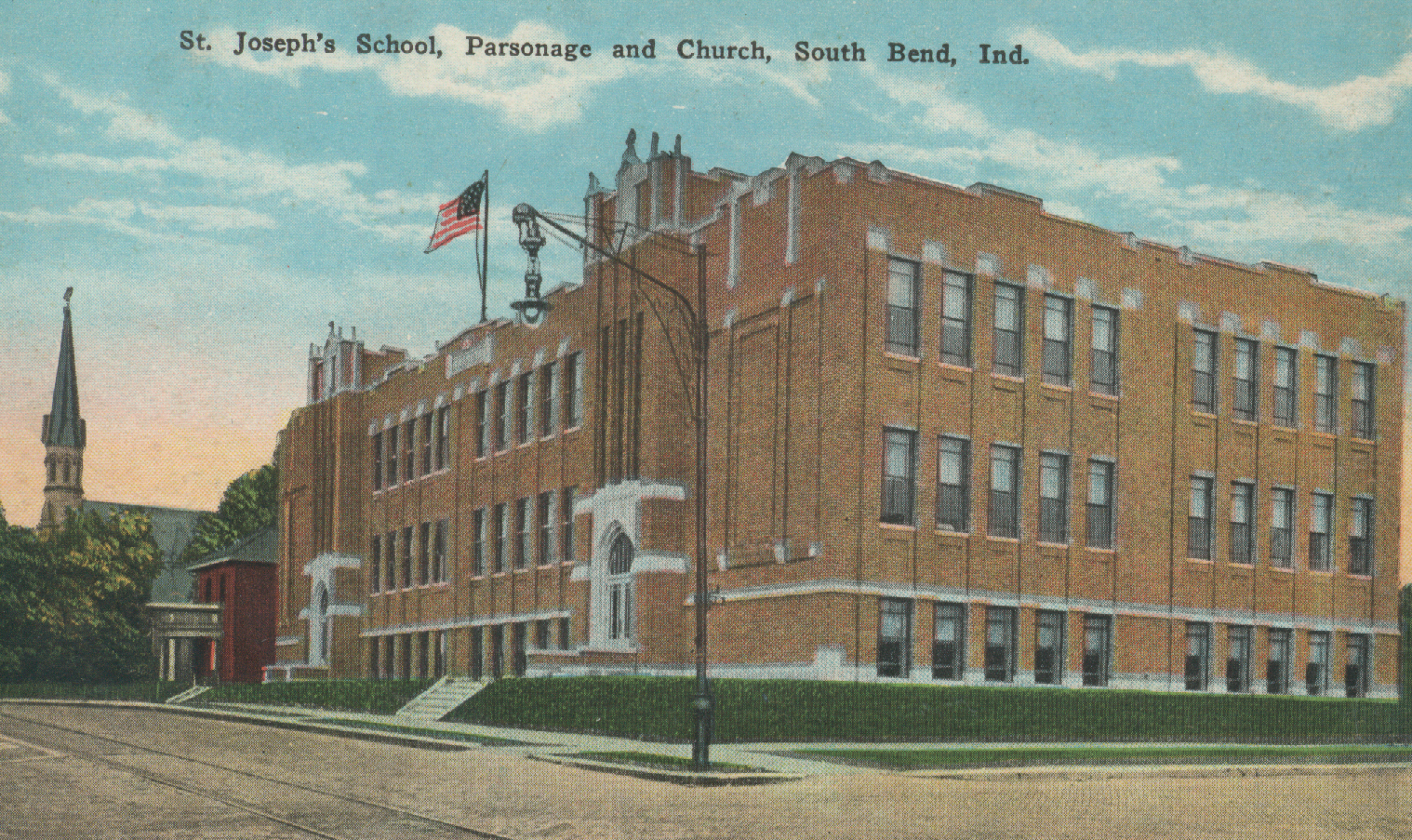 Historical photograph of St. Joseph's School, Parsonage, and Church in South Bend, Indiana, showing a large brick school building with multiple windows, an American flag, and a church steeple in the background under a partly cloudy sky.