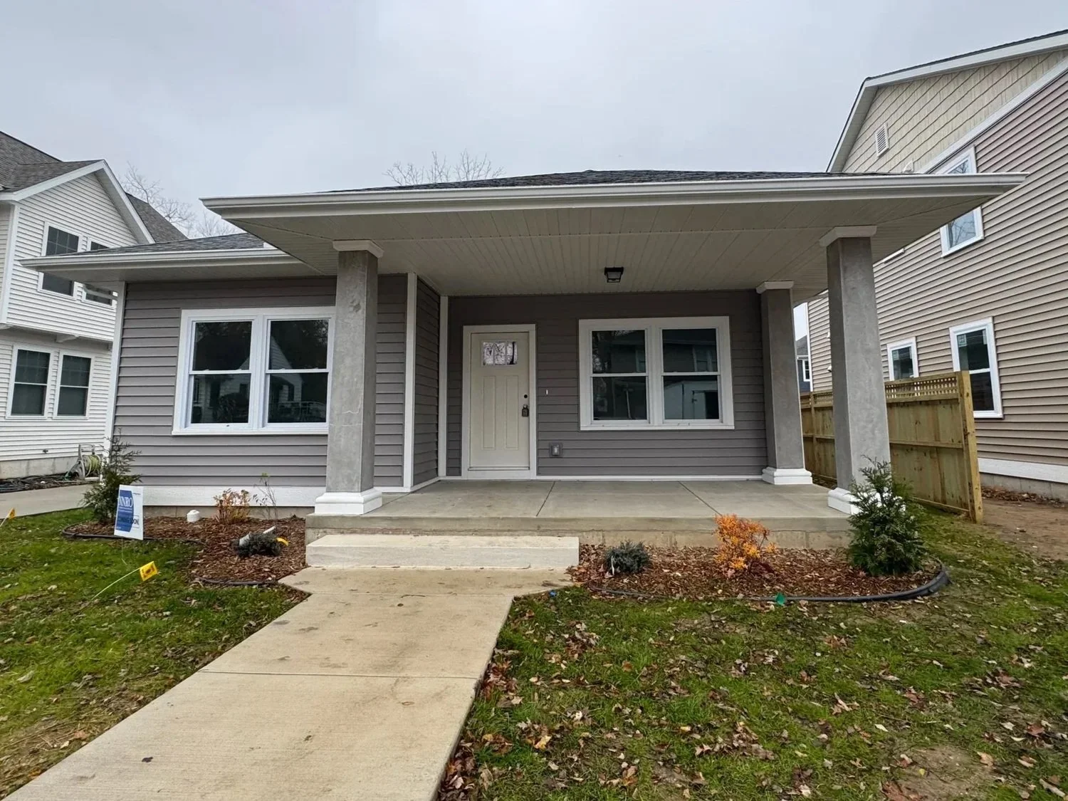 Newly constructed small house with a front porch, two windows, gray siding, and a white front door, surrounded by a lawn with small plants and a sidewalk leading up to the porch.