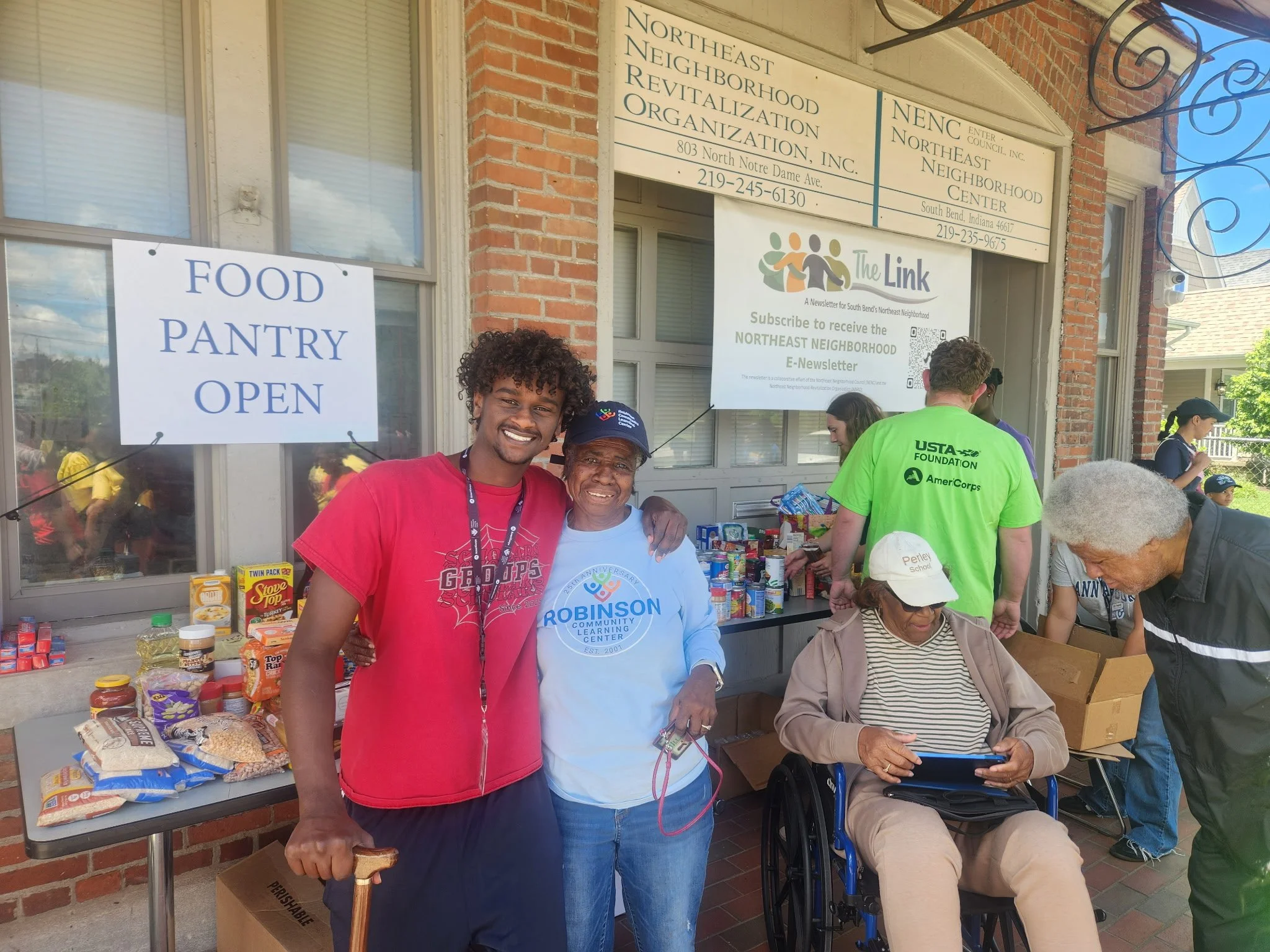 People gathered outside a building with signs for a food pantry and community organization, with boxes of food on a table. Two women in the foreground smiling, one with a cane, others in the background engaged in activities.