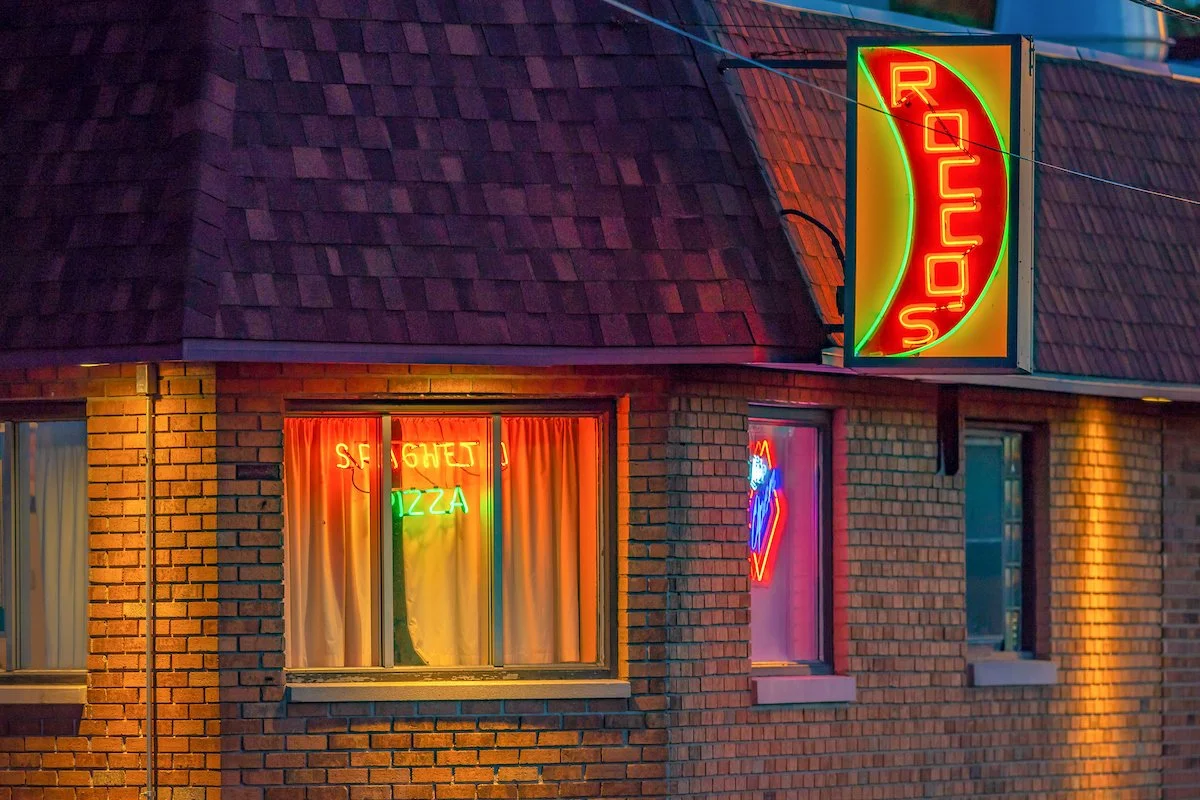 Exterior of a brick building with neon signs advertising a 'ROUSE' and 'STEAK AND PIZZA,' illuminated in red, green, and blue colors. The building has a shingled roof, and the windows display curtains and neon signs.