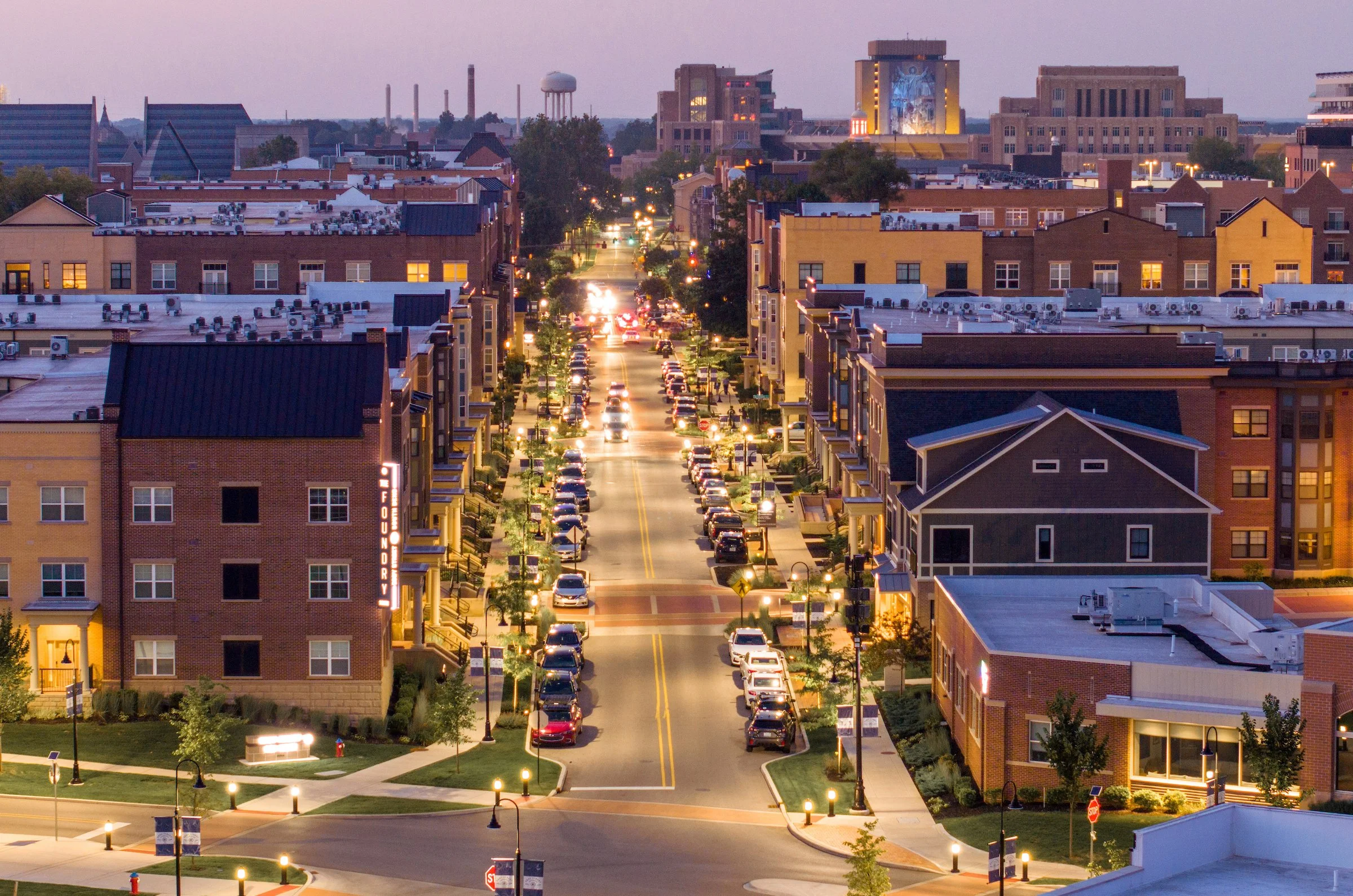 A cityscape view at dusk showing a street with parked cars, apartment buildings, and a skyline with taller buildings and smokestacks in the background.