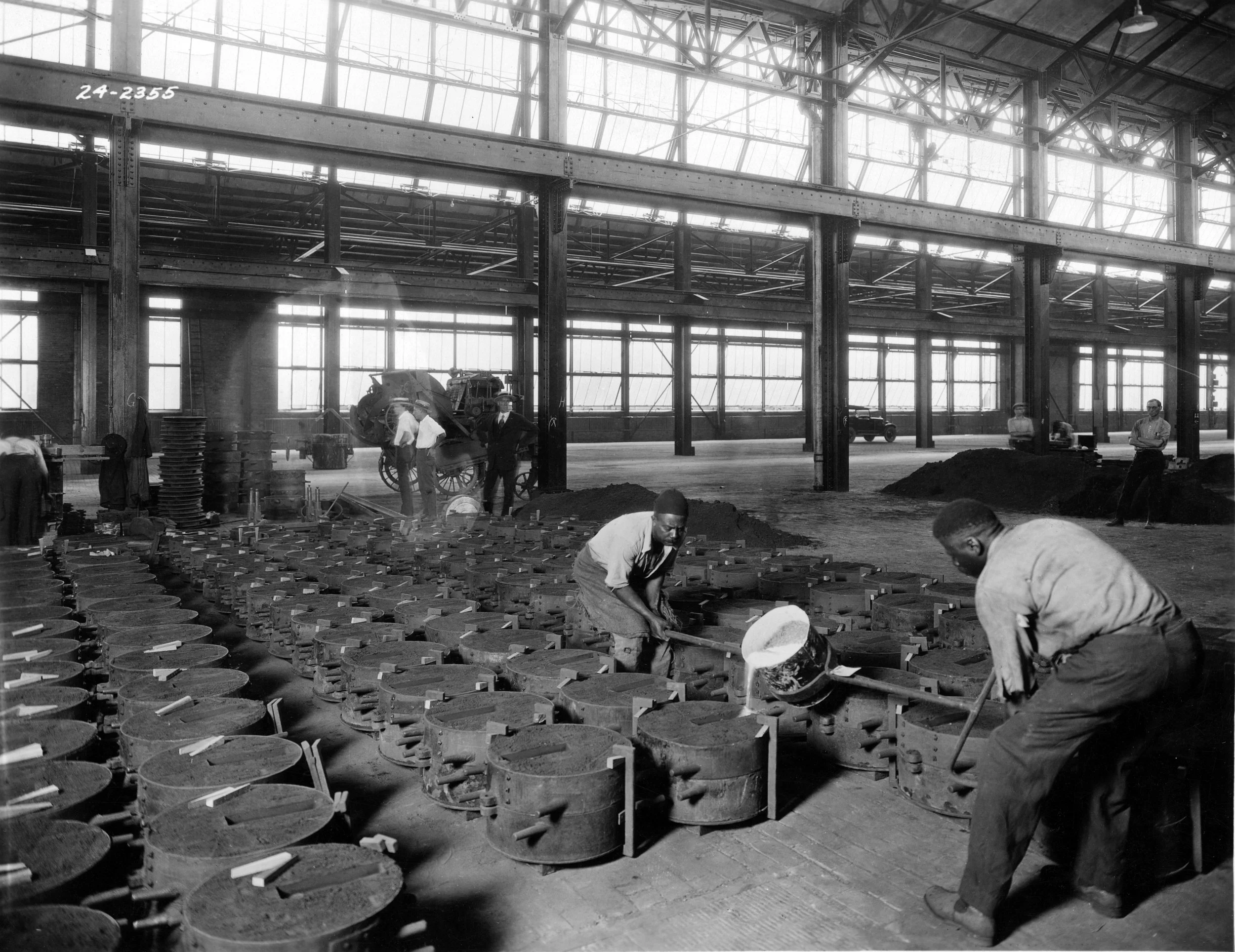 Black and white photo of workers inside a large industrial building working on metal parts arranged in organized rows with some workers bending over and others standing.