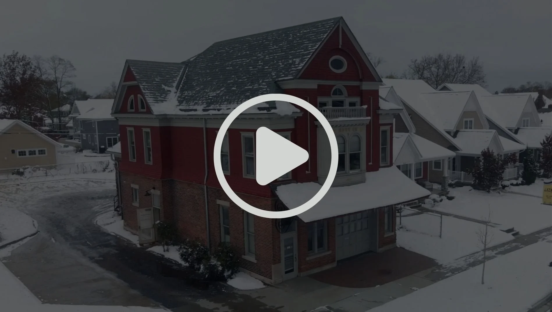 A multi-story red Victorian-style house with a front porch, surrounded by snow-covered lawns and neighboring houses, under a cloudy sky.