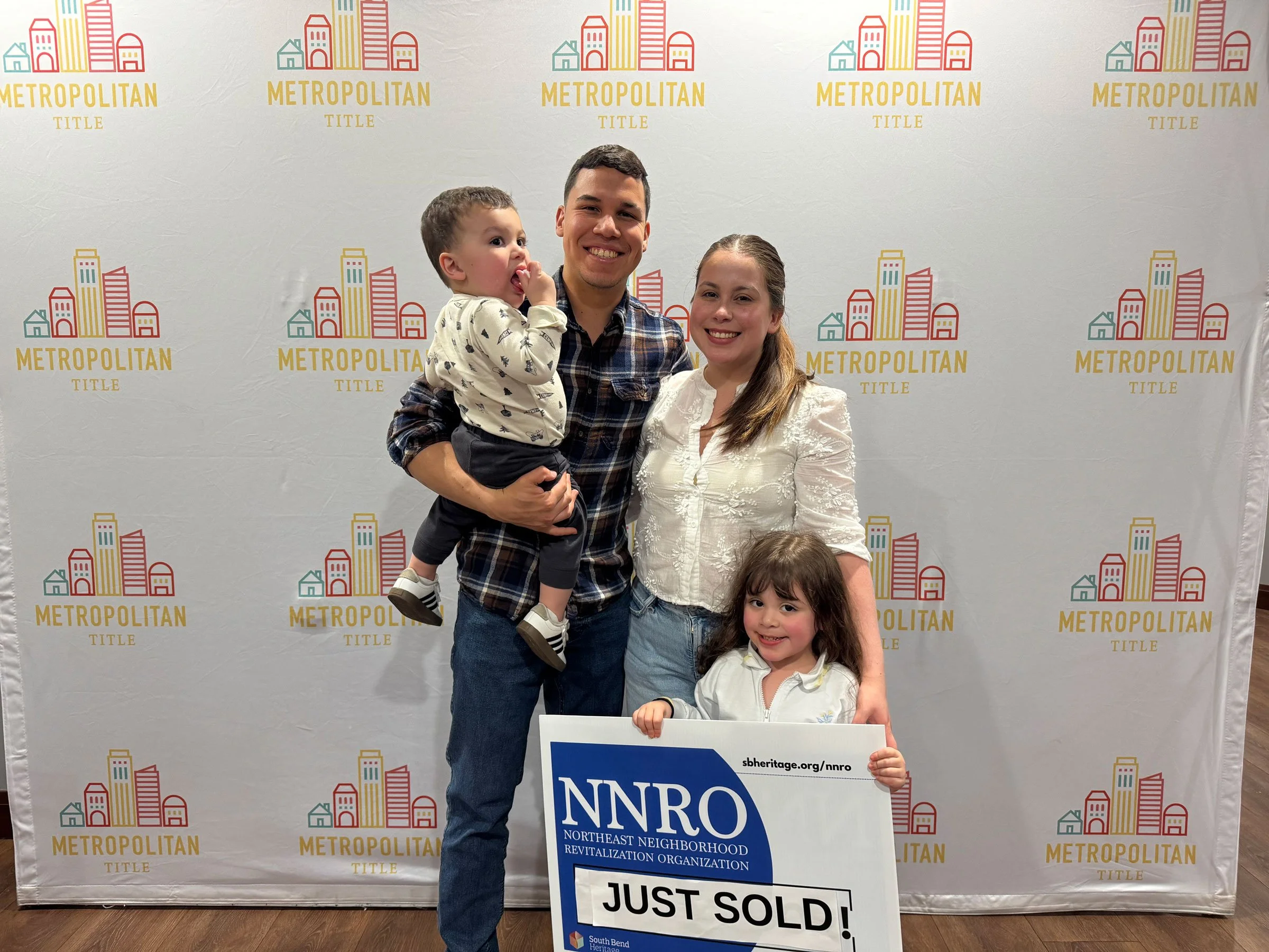 A family of four, a man, a woman, and two young girls, standing in front of a backdrop with the words METROPOLITAN TITLE and a colorful cityscape drawing. The man is holding a young boy, and the woman is holding a sign that reads 'NNRO' and 'JUST SOLD!'