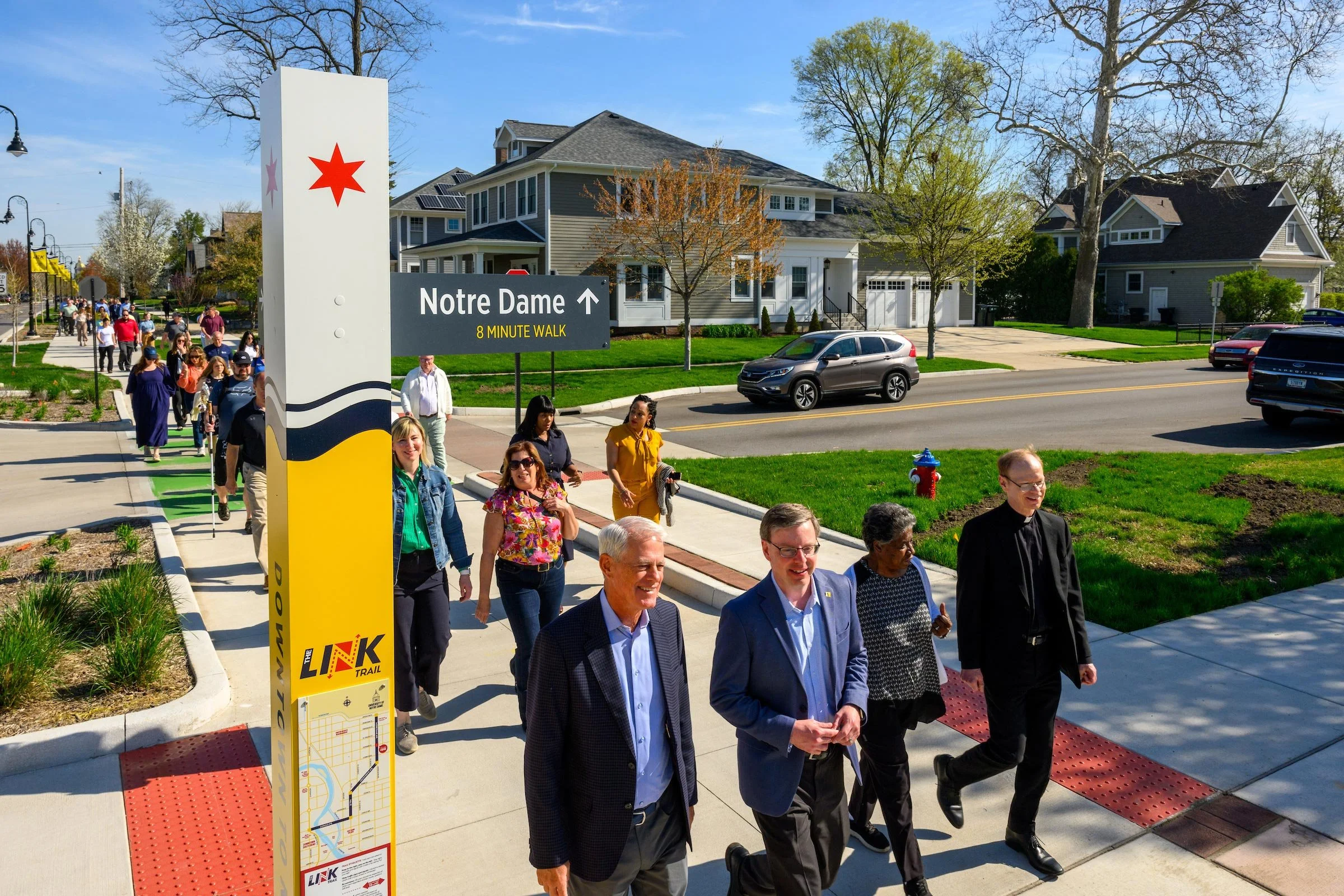A group of people walking on a sidewalk along a street with houses in the background. There is a sign that reads 'Notre Dame 8 Minute Walk' with an arrow pointing straight ahead.