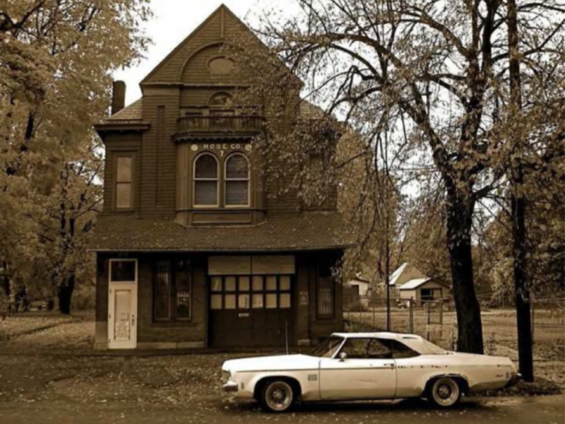 An old Victorian-style building with a sign that reads "HOSC CO." in the upper floor window, and a vintage white car parked in front, surrounded by trees with autumn leaves.