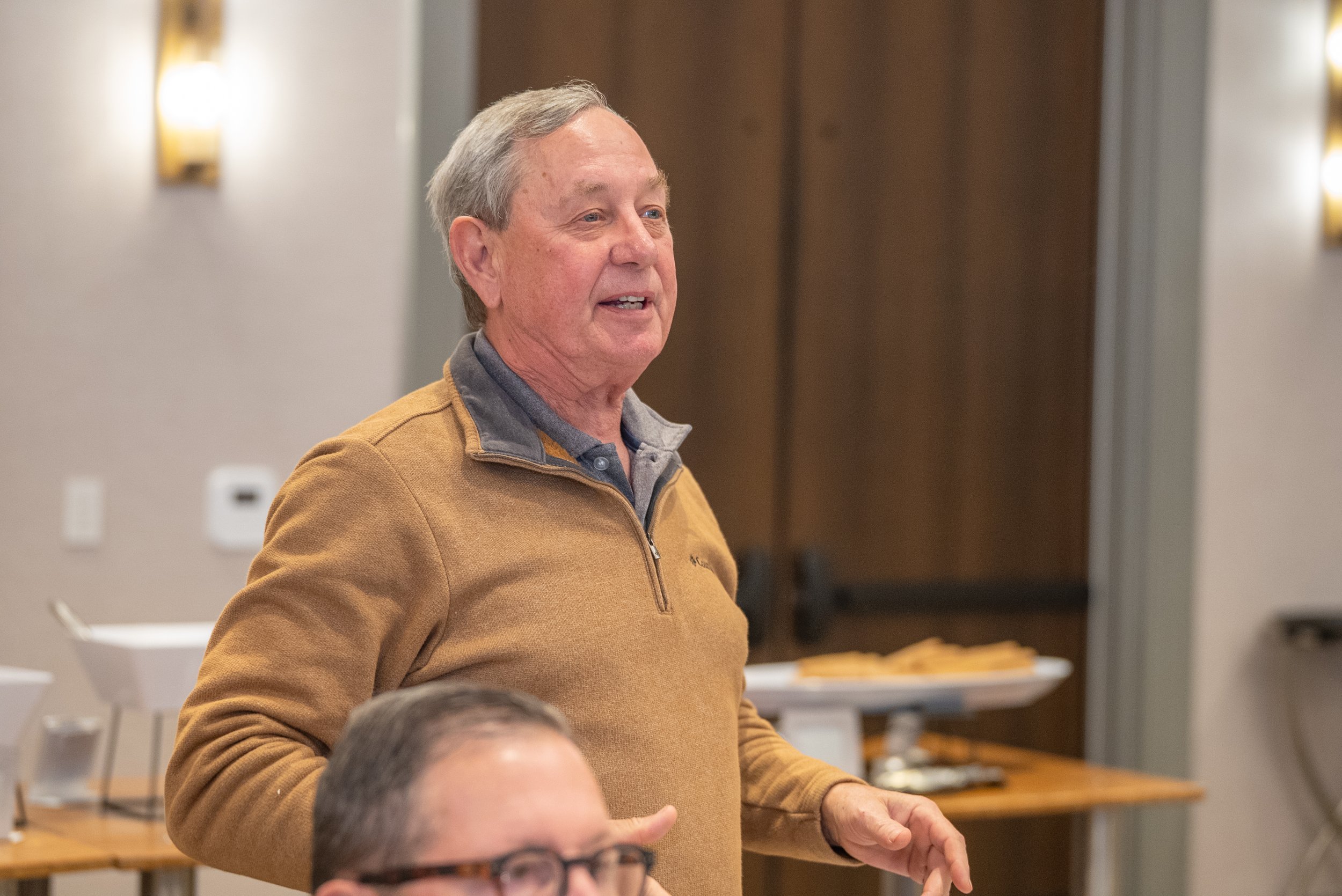 An elderly man with gray hair speaking or listening at an indoor gathering, wearing a light brown quarter-zip sweater with a gray collar.