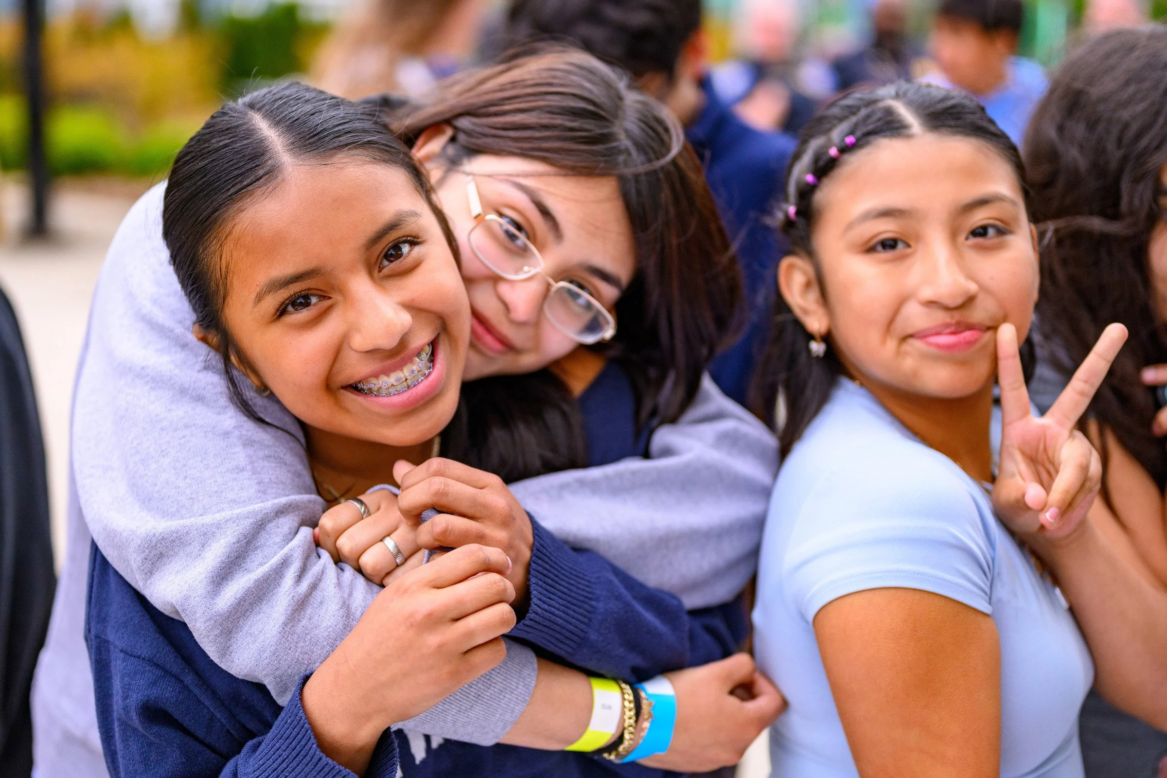 Group of young girls hugging and smiling at an outdoor event, with some making peace signs and wearing colorful bracelets.