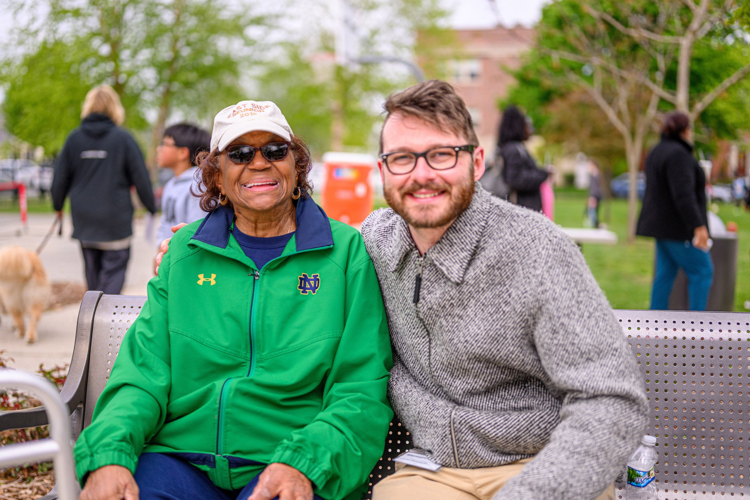 A smiling elderly woman wearing sunglasses, a beige cap, and a green jacket sitting beside a smiling young man with glasses, a beard, and a gray patterned zip-up jacket at an outdoor park event with trees, other people, and a water cooler in the background.