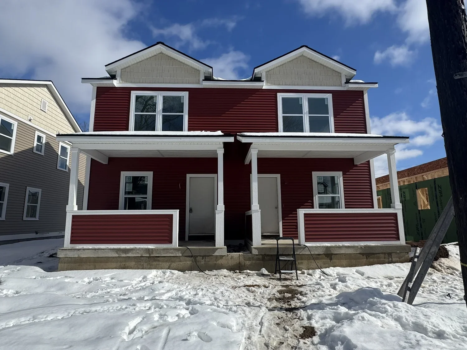 A partially constructed two-story house with red vinyl siding and white trim, snow on the ground, and a blue sky with some clouds.