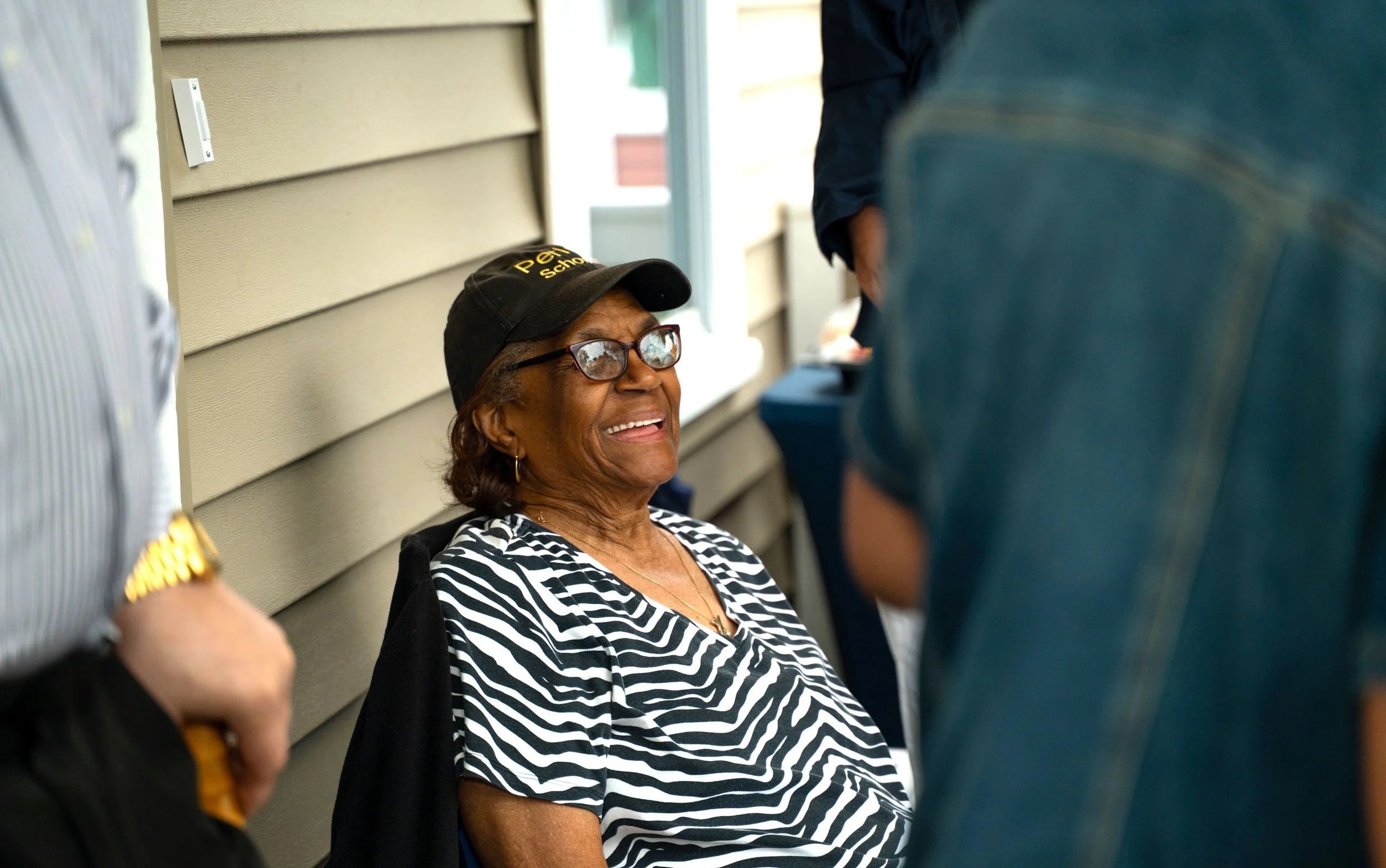 An elderly woman wearing glasses and a black cap with yellow writing, sitting outside near a beige wall, smiling and talking to someone.