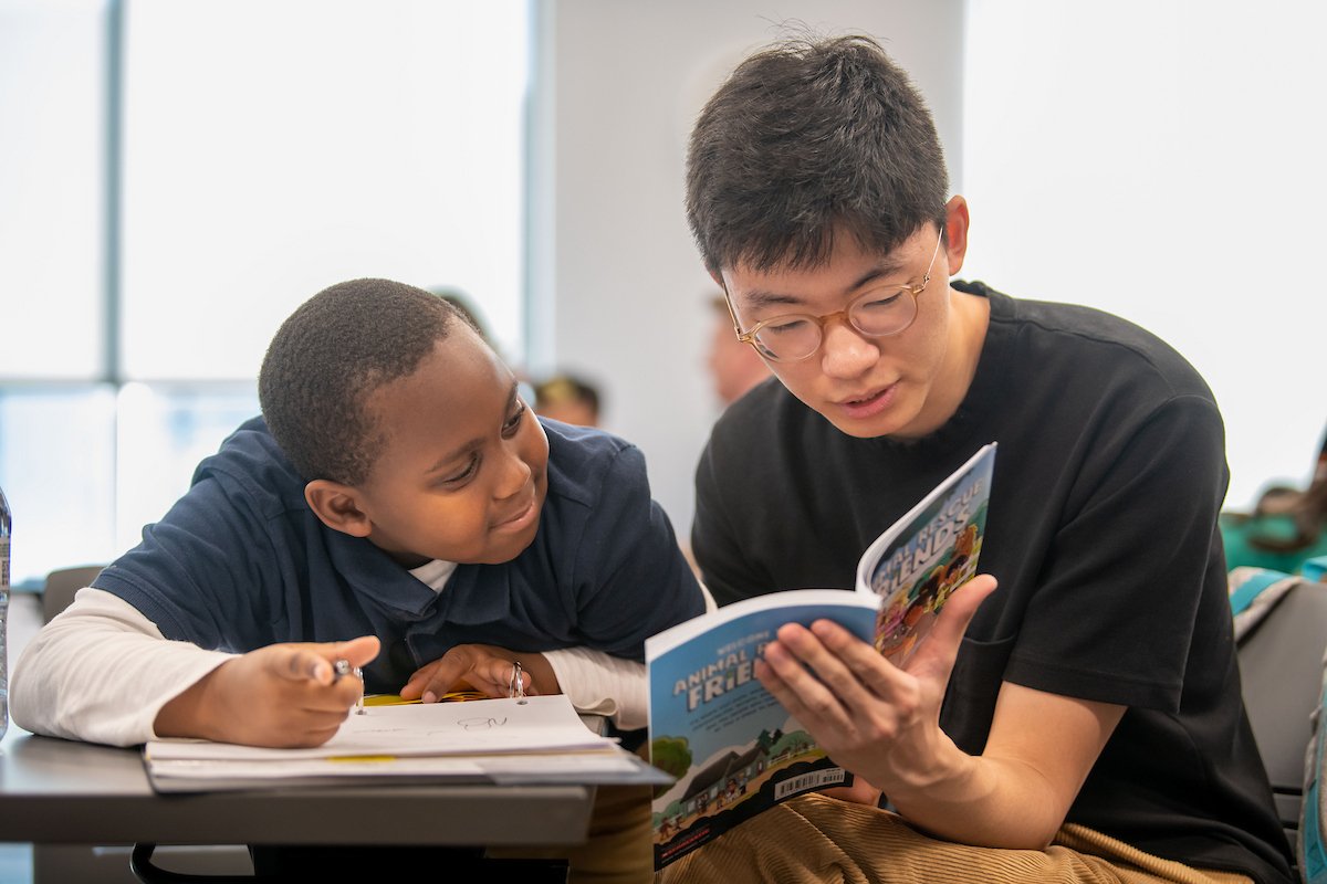 A young boy and an adult male reading a comic book together at a table in a classroom or library setting.