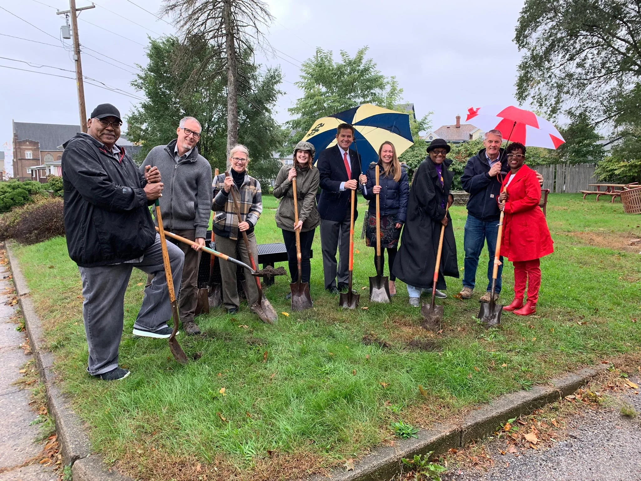 A group of people standing outdoors in light rain on grass, holding shovels, with some holding umbrellas, smiling for a photo during a community event in a neighborhood with trees and houses in the background.