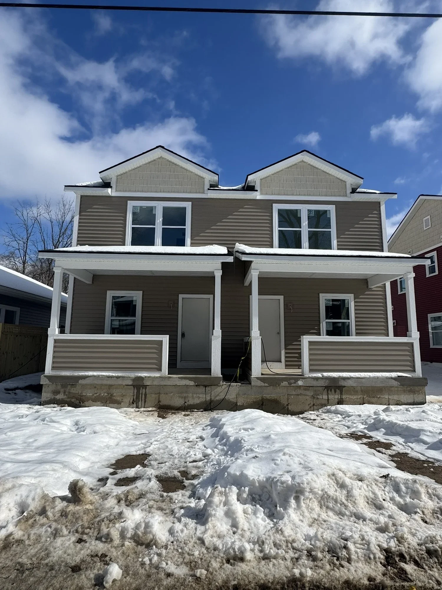 Front view of a two-story house with beige and white siding, snow on the porch roof and ground, and a partly cloudy blue sky above.