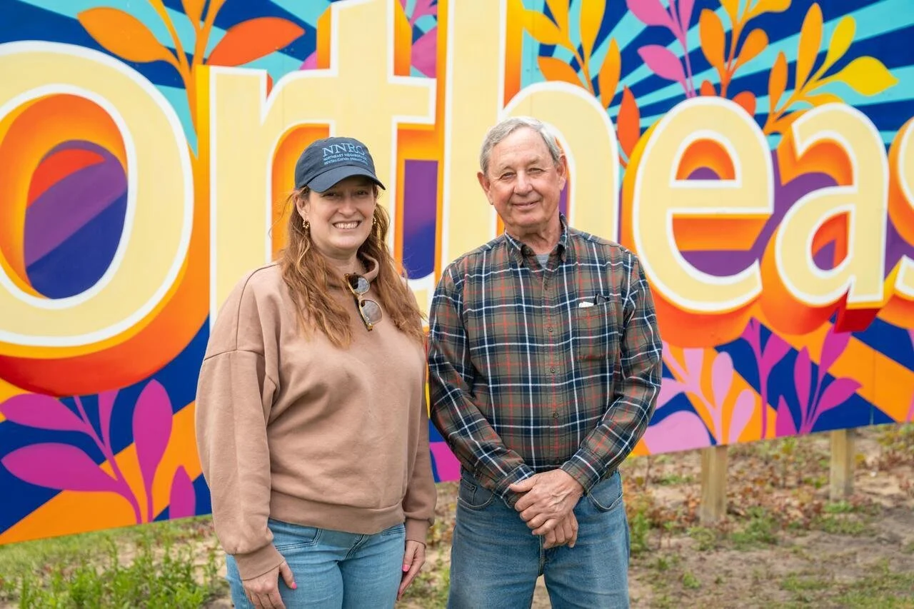Two smiling people, a woman with long red hair, wearing a beige sweatshirt, jeans, and a blue cap, and a man with gray hair, wearing a plaid shirt, stand in front of a colorful sign that says 'Orleans' with floral decorations.